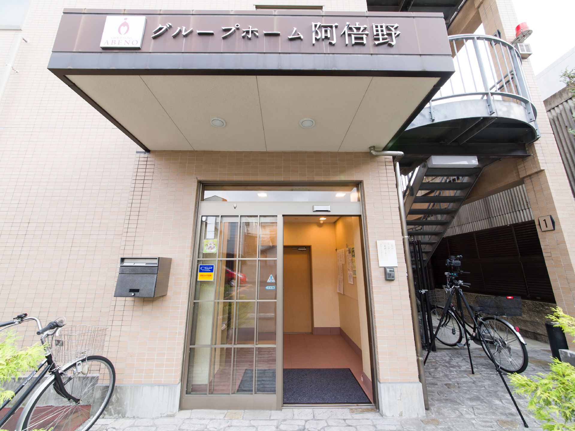 施設の玄関ドアの上部には大きな四角い屋根が設置されている。前面はブラウンで、シルバーの文字で施設名が書かれている。
