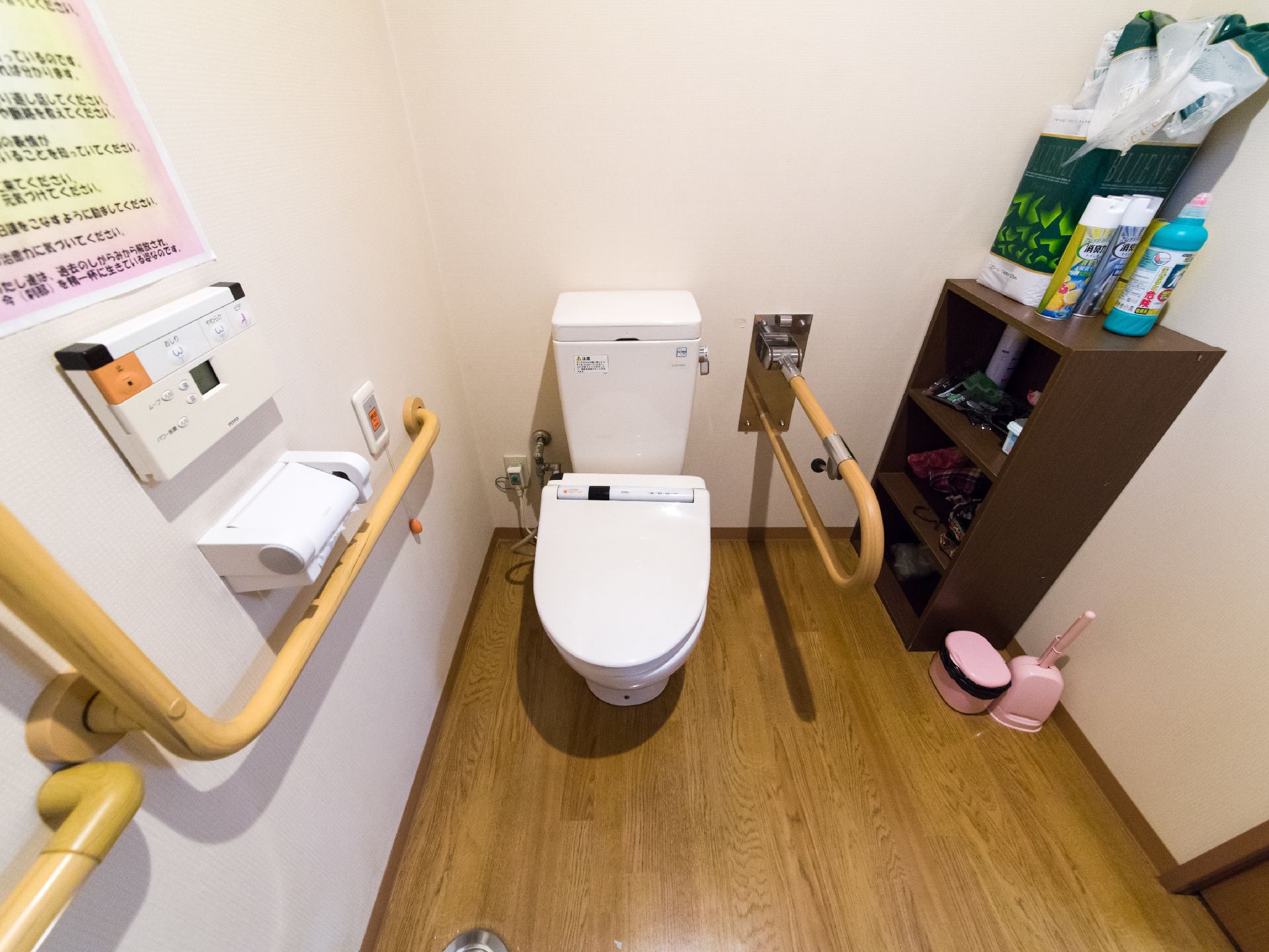 白色と木目仕様のトイレスペースで便器の両側には手すりが取り付けてある。また隣には焦げ茶色の3段ボックスが置かれている。