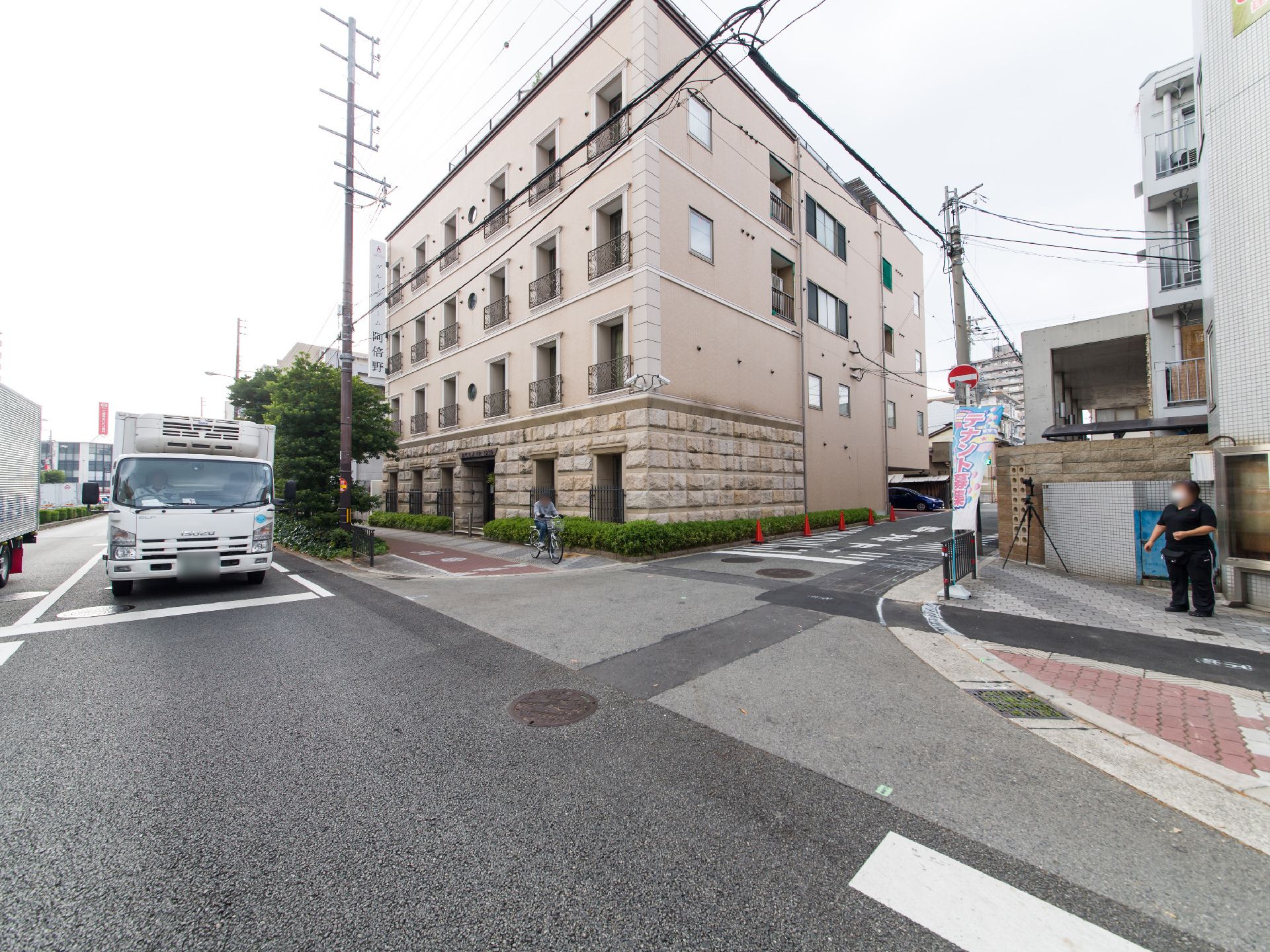 施設の建物は道路の曲がり角部分に建っている。外壁の色はベージュ系で、窓の外側には黒いフェンスが設置されている。