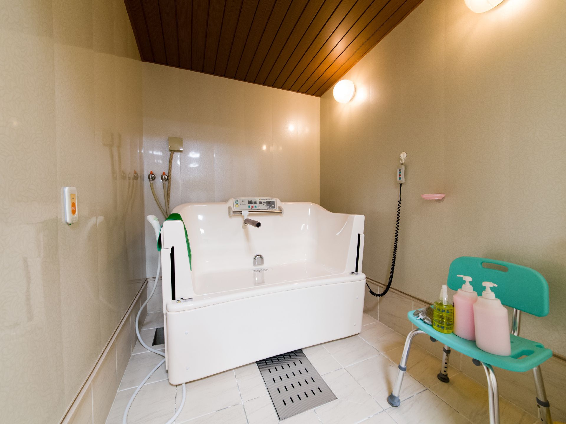 洗浄を介助するための大型器具がある。壁にリモコンが吊り下げられている。浴室用のイスにボトルが3つ置いてある。