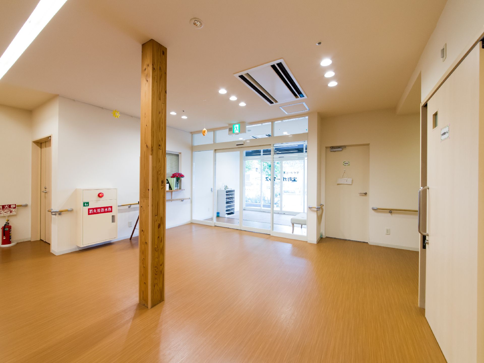木目調の柱が1本ある。出入り口付近には窓口があり、赤い花が飾られている。ガラスのドアには施設名が見える。