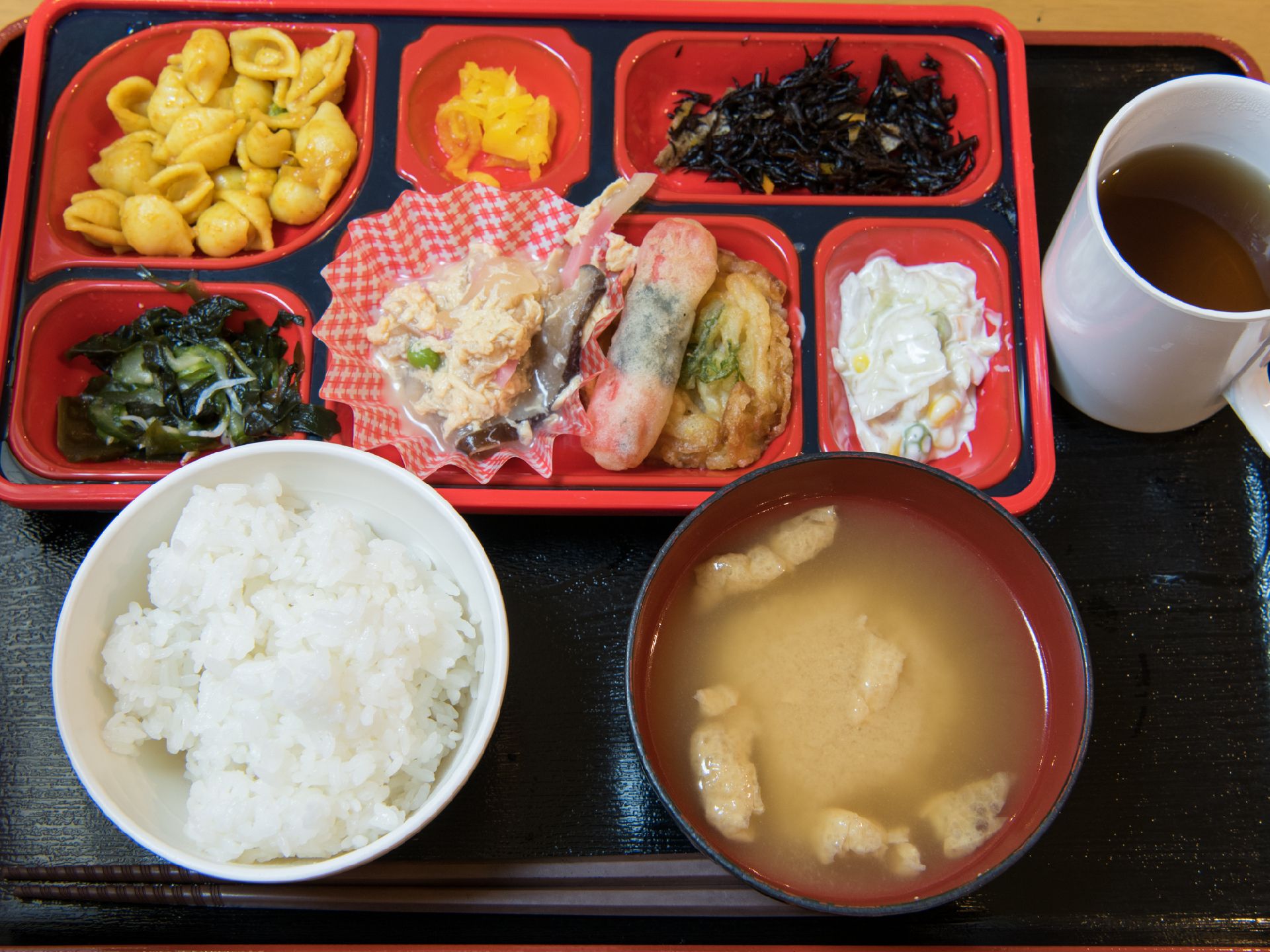 ご飯、味噌汁、おかずプレート、お茶の食事。おかずは、サラダ、酢の物、漬物、煮物などバラエティに富んでいる。