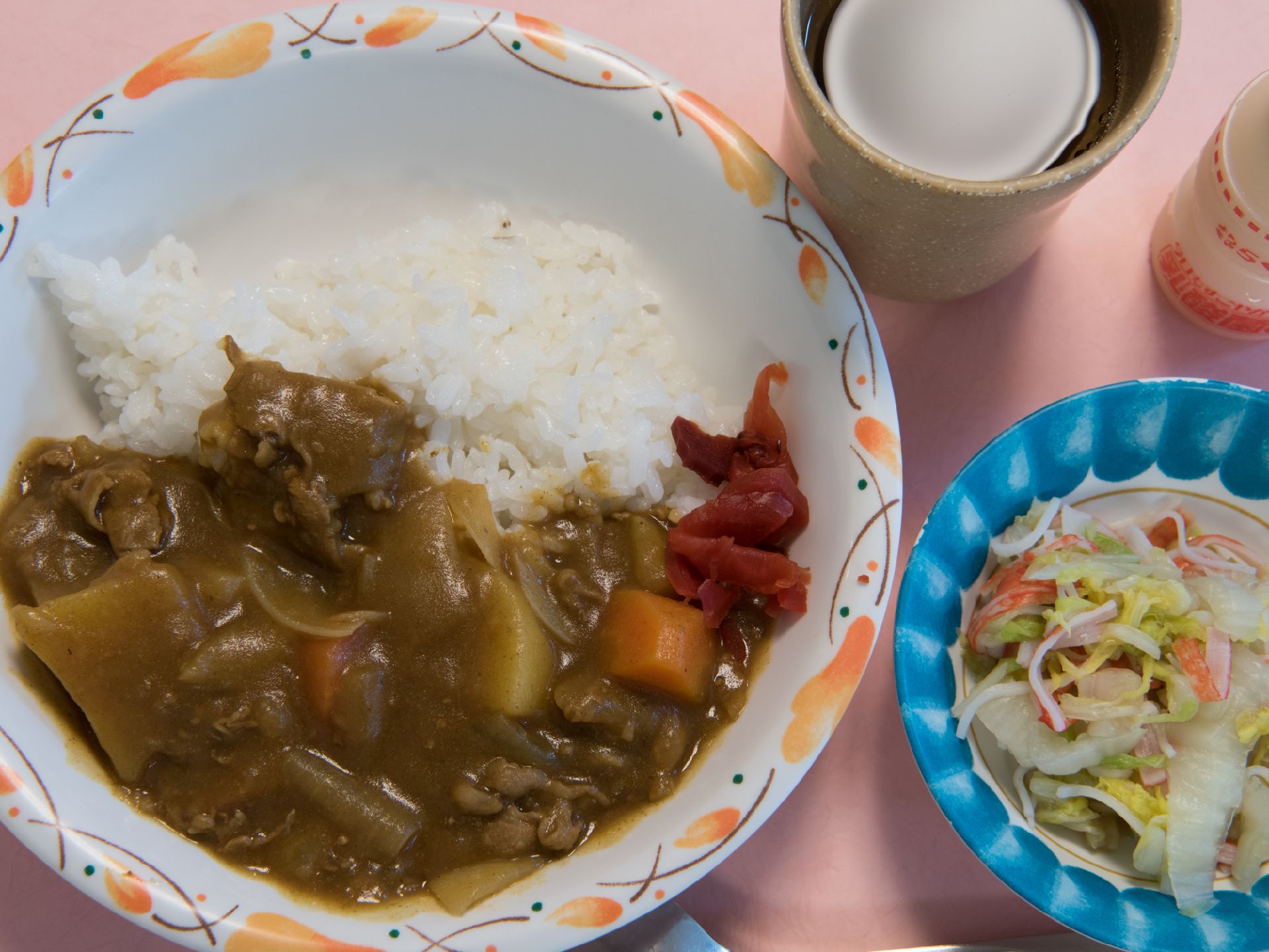 福神漬けの添えられたカレーライスにカニかまぼこと白菜の和え物。湯呑に入ったお茶と、乳酸菌飲料も出されている。