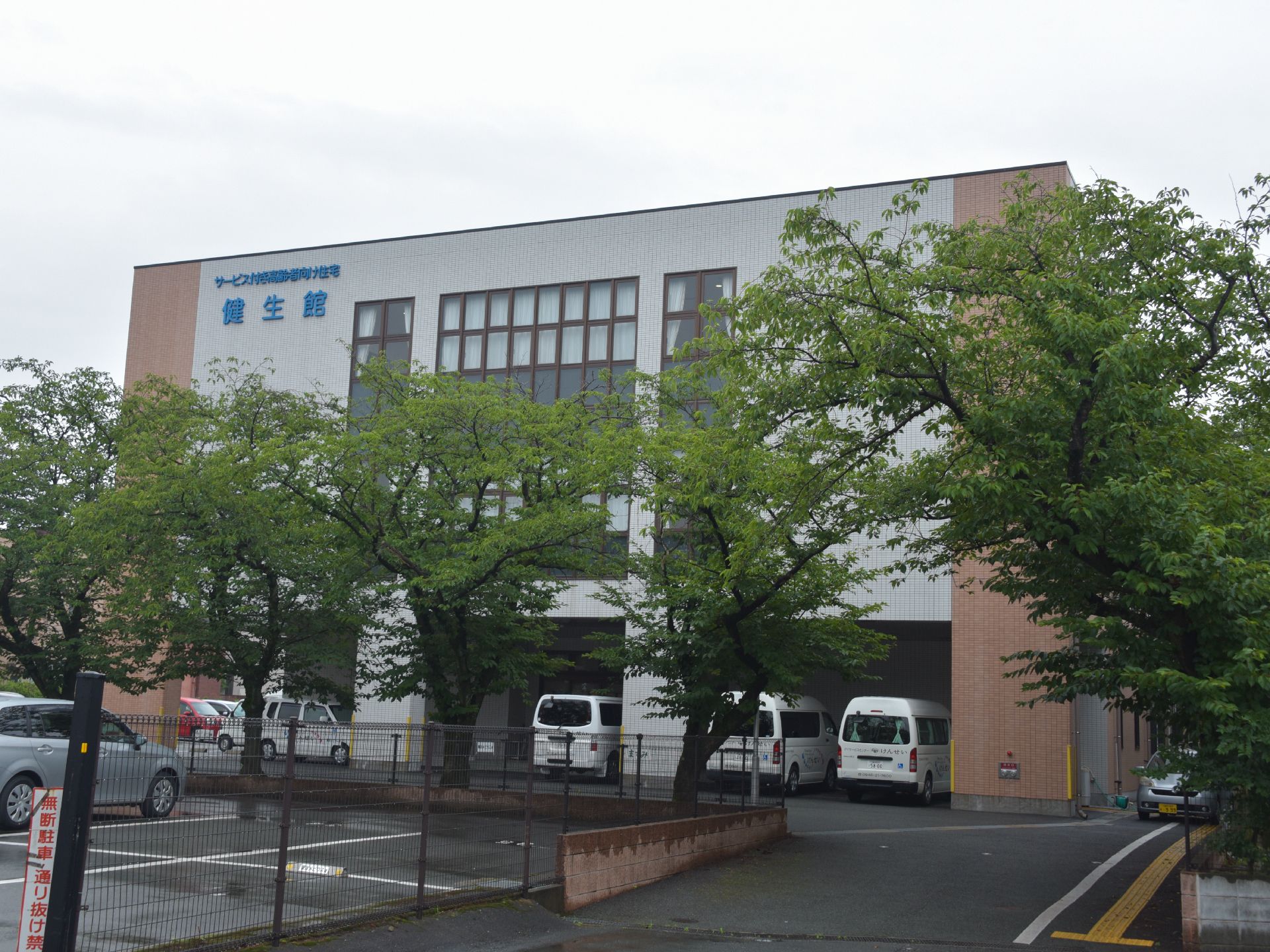 サービス付き高齢者向け住宅、健生館の外観は、玄関側に緑の葉が茂る木が出迎え、落ち着いた雰囲気。雨の日は車から玄関まで濡れない作りである。
