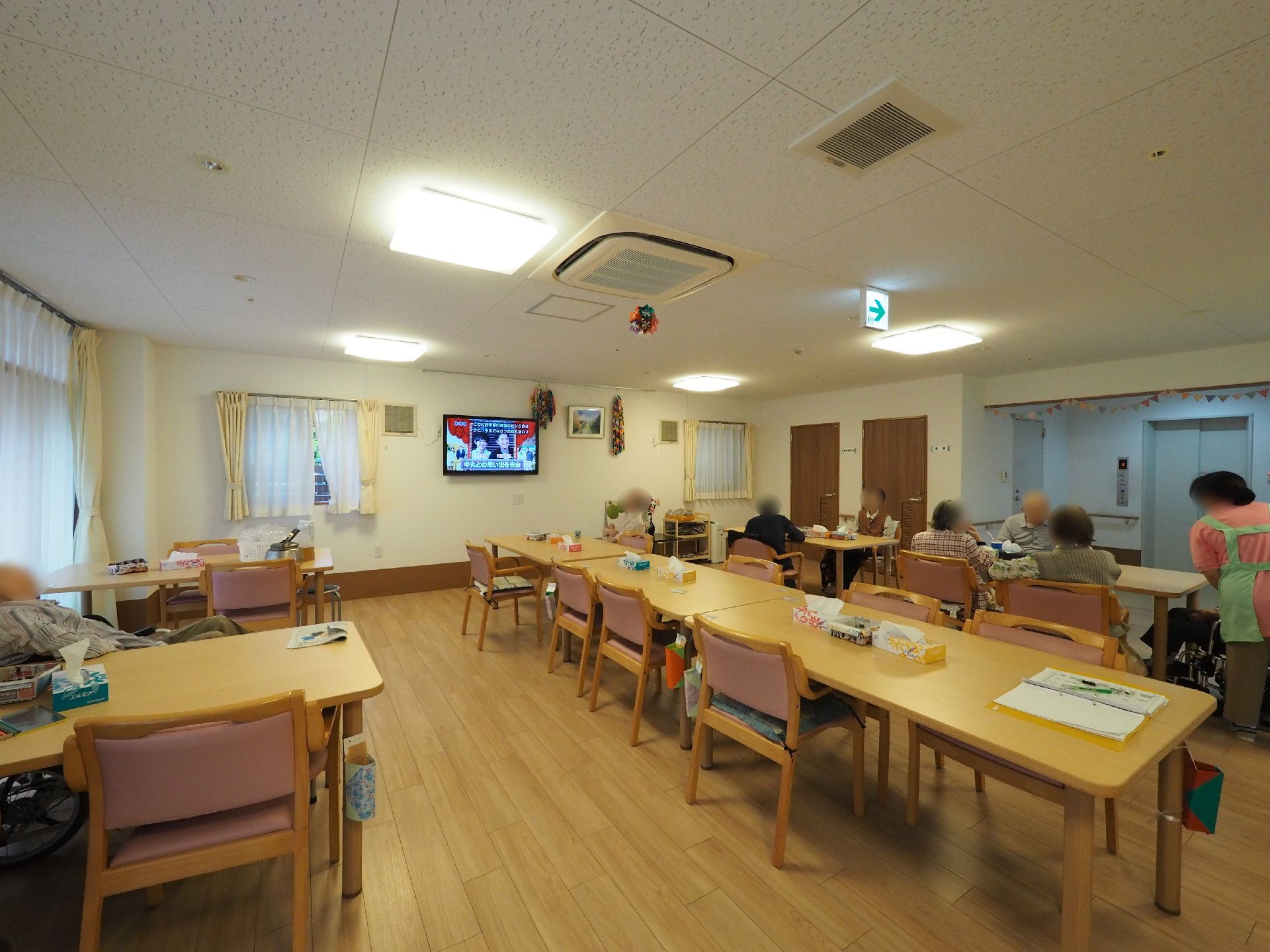 広い食堂にはテーブルが並べて設置してある。入居者やスタッフがくつろいでいる様子。壁には大きな液晶テレビがある。