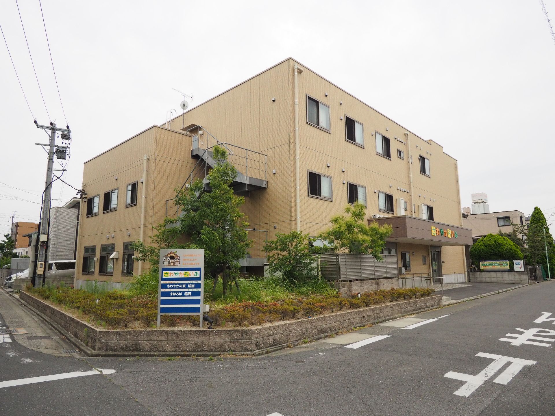 施設外観の様子。タイル張りのベージュ色の建物で、道路に面したところに看板が設置されている。非常階段が見えている。