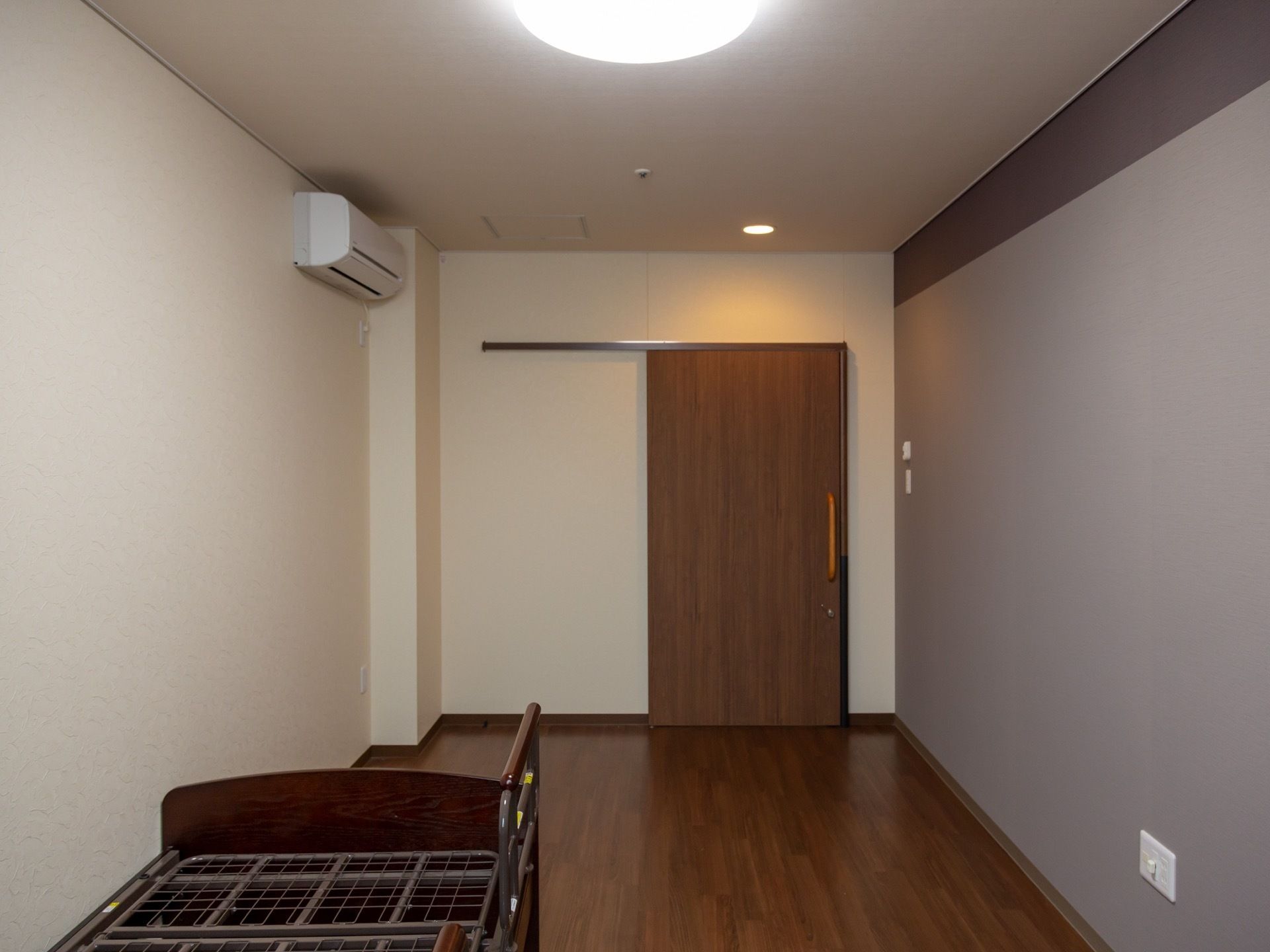居室は落ち着いた色合いのフローリング張りとなっている。介護用ベッドとエアコン、照明器具が設置されている。