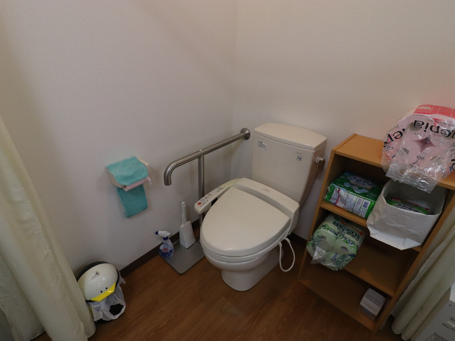 カーテンで仕切られるタイプのシャワー付き洋式トイレ。トイレの横に棚があり、介護用おむつやトイレットペーパーが置いてある。