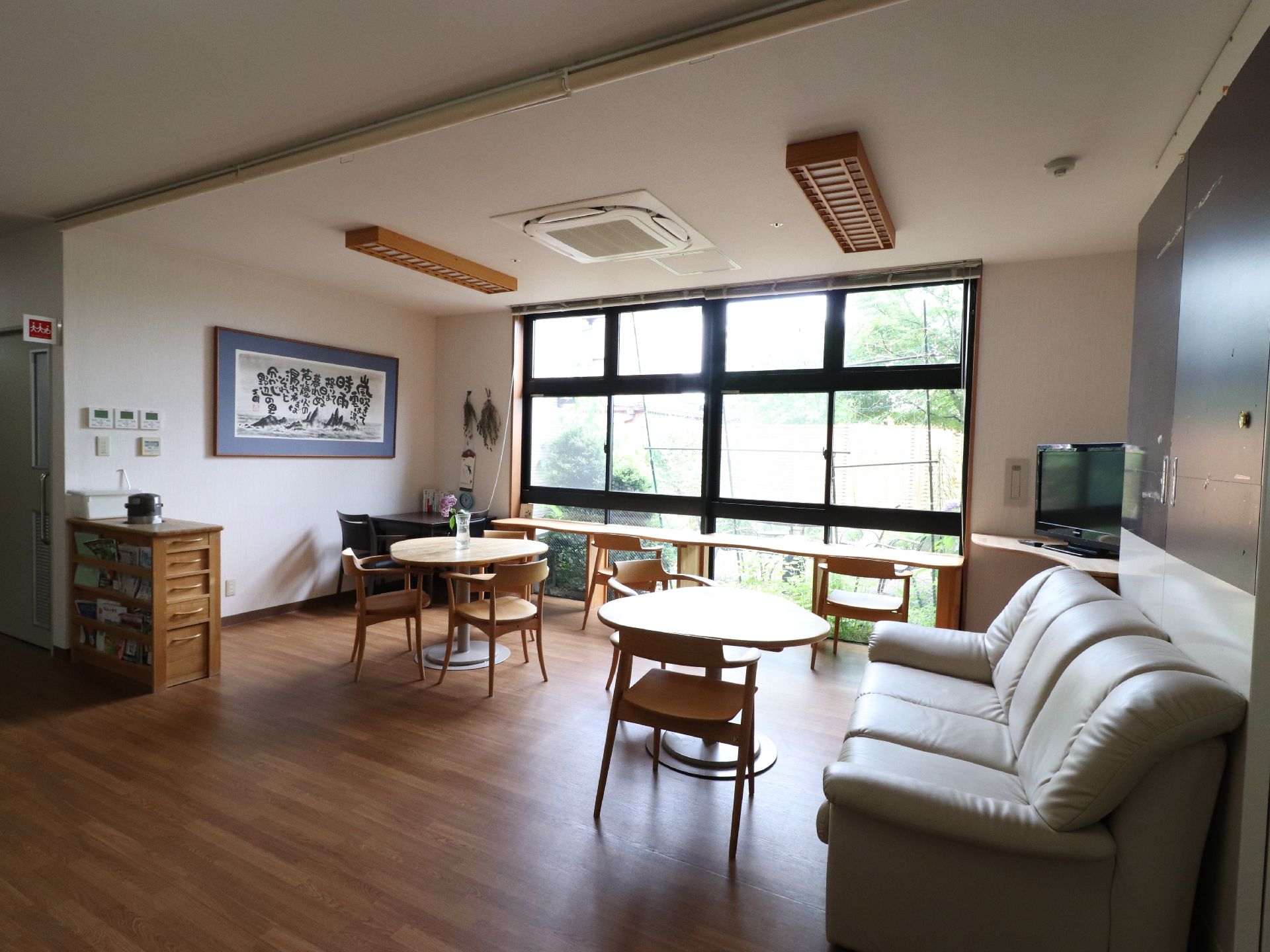 窓側に長テーブルがあり、手前に丸テーブルが二つ配置されている。角にテレビがある。ソファは3人掛けである。