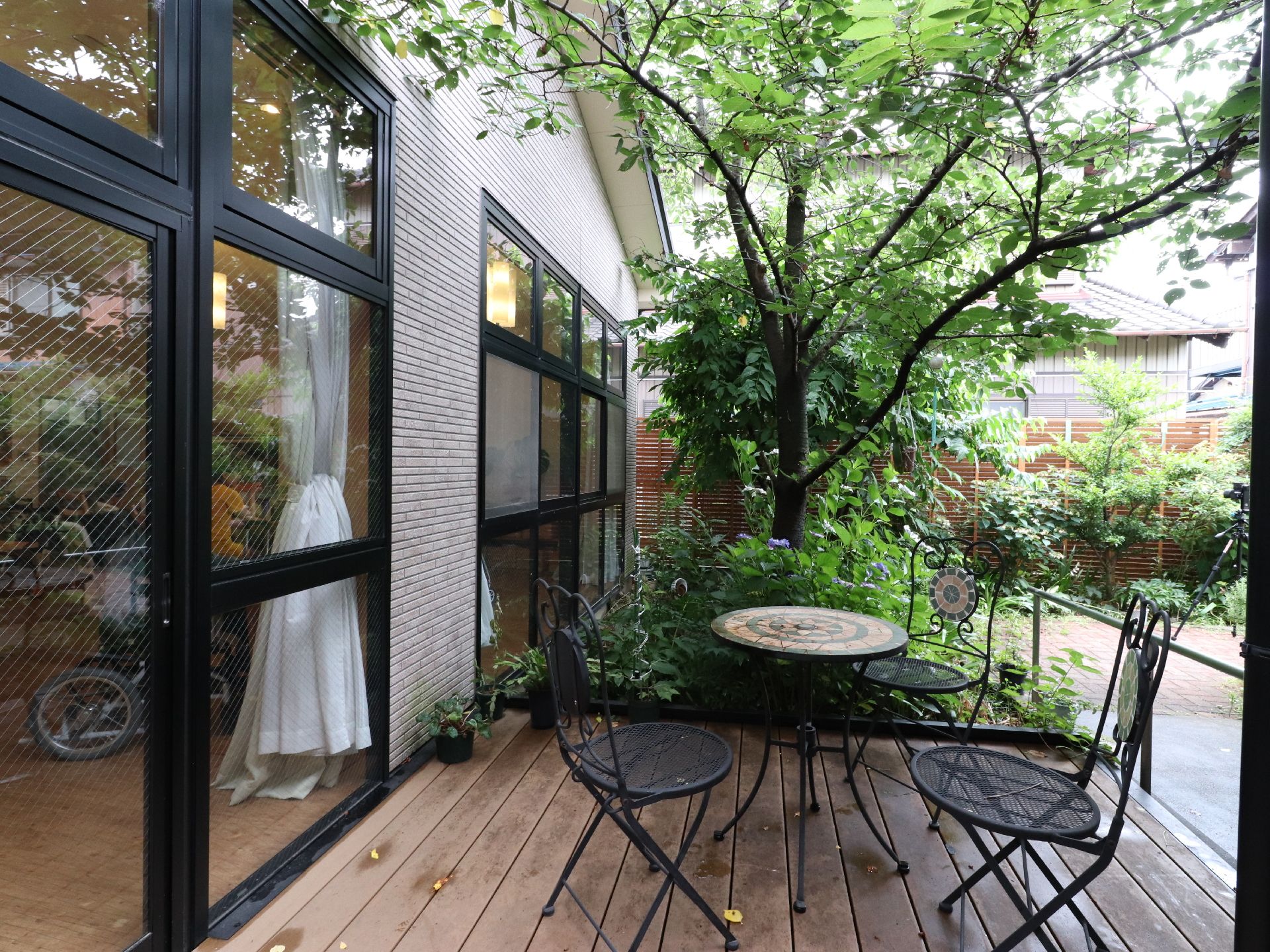 ウッドデッキには庭用のテーブルといすが置いてある。庭には桜の木のほか、いろいろな木が植えてある。デッキの外側には手すりがついている。