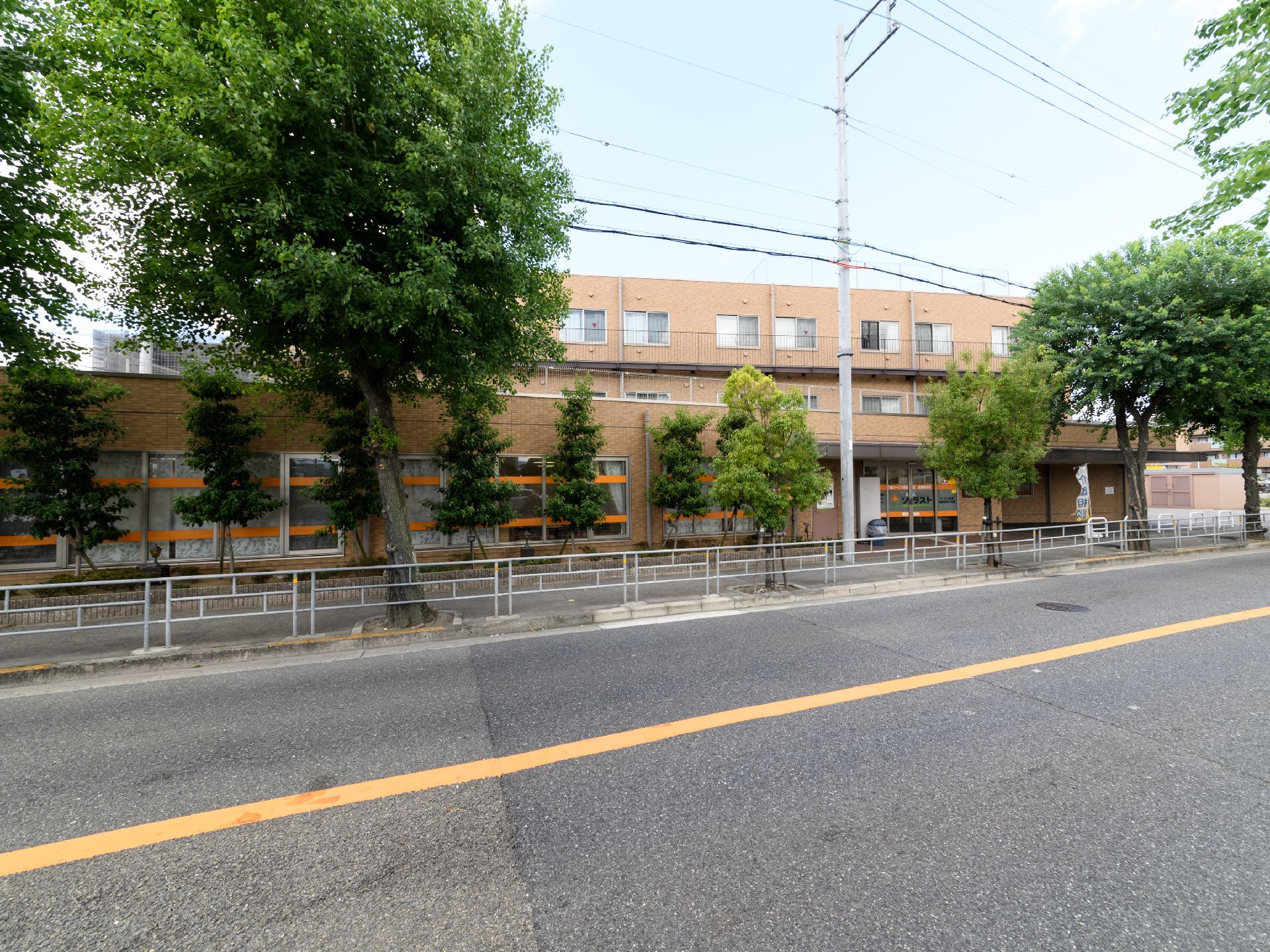 車道を挟んだ施設の外観の様子。3階建ての建物で、1階部分にはオレンジ色のラインの入った窓が並んでいる。施設の周りには街路樹がある。