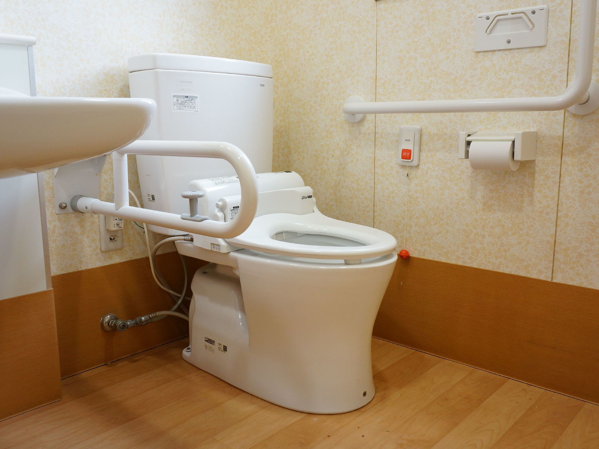 壁には緊急時呼び出しブザーや手すりが設置されている。便器脇にも可動式の手すりがある。個室内に洗面台がある。