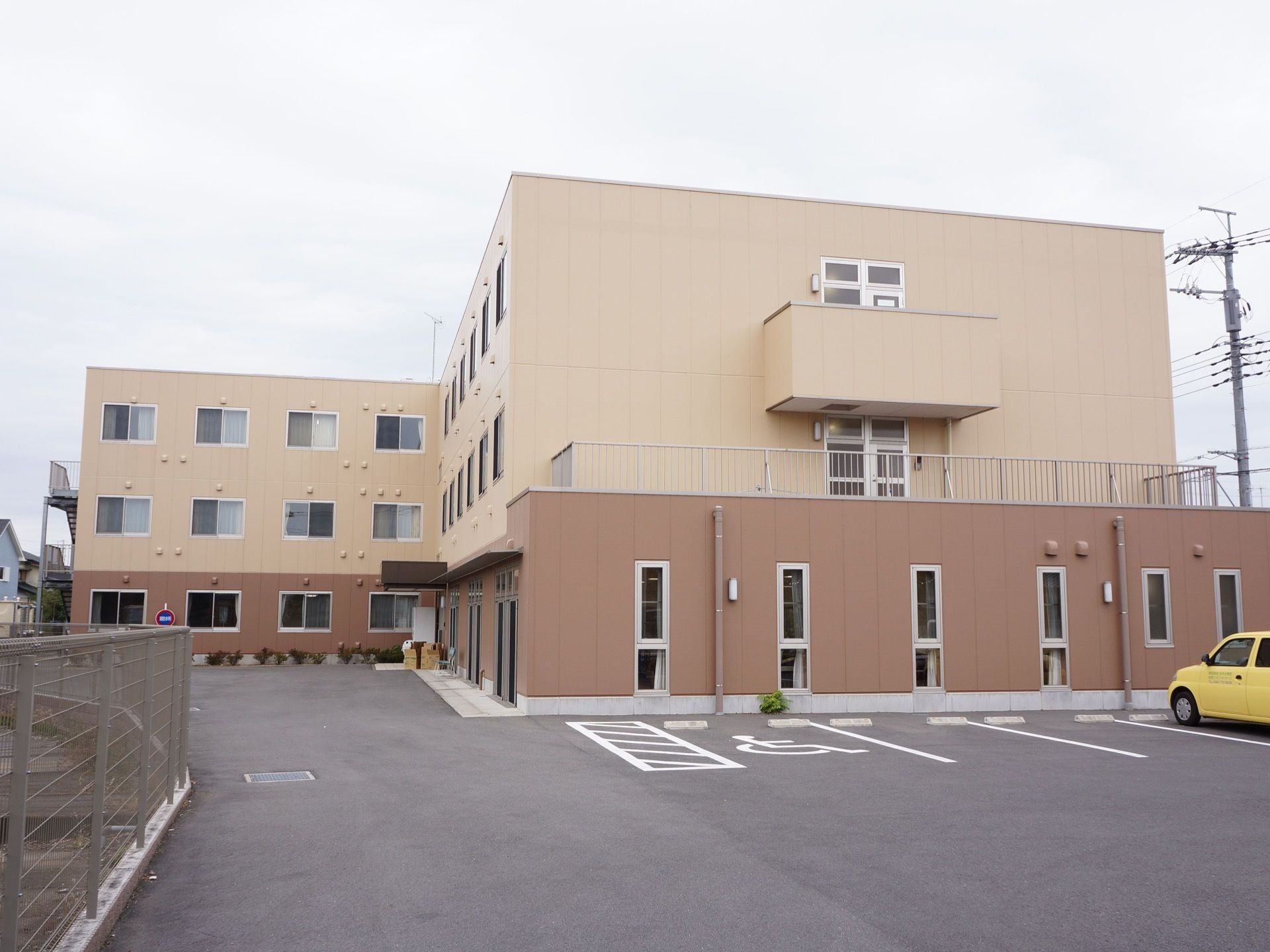 施設の建物は二階建てで、外壁は明るいブラウンの濃淡でデザインしている。アスファルト舗装の駐車場を完備している。