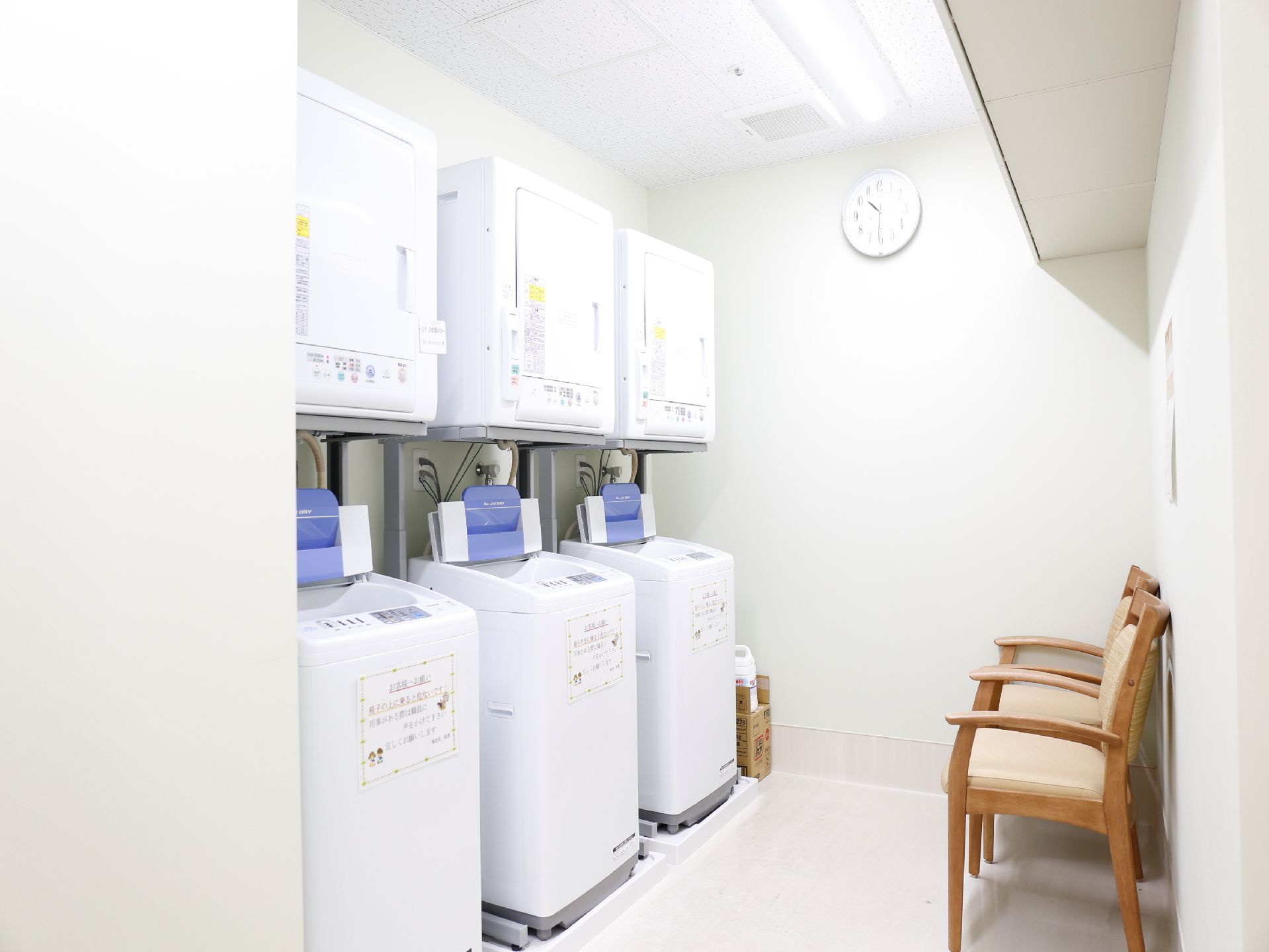 白で統一された部屋に時計がかかっている。3台の洗濯機と乾燥機があり、手前には3脚の椅子が置かれている。