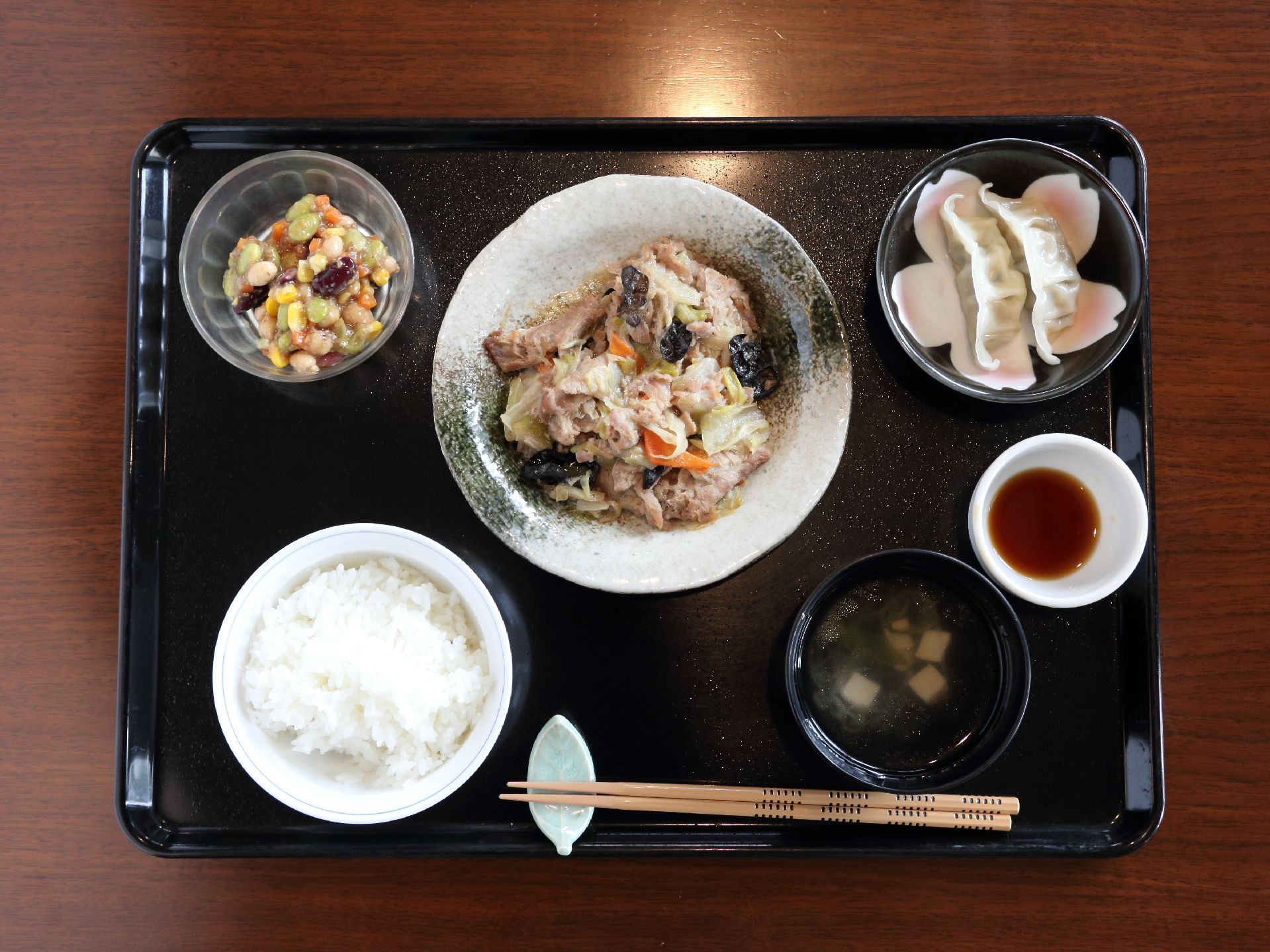 黒いお盆に食事が乗せられている。ご飯、味噌汁の他、中央にメインのおかず、小鉢、餃子がある。手前に箸置きもセット。