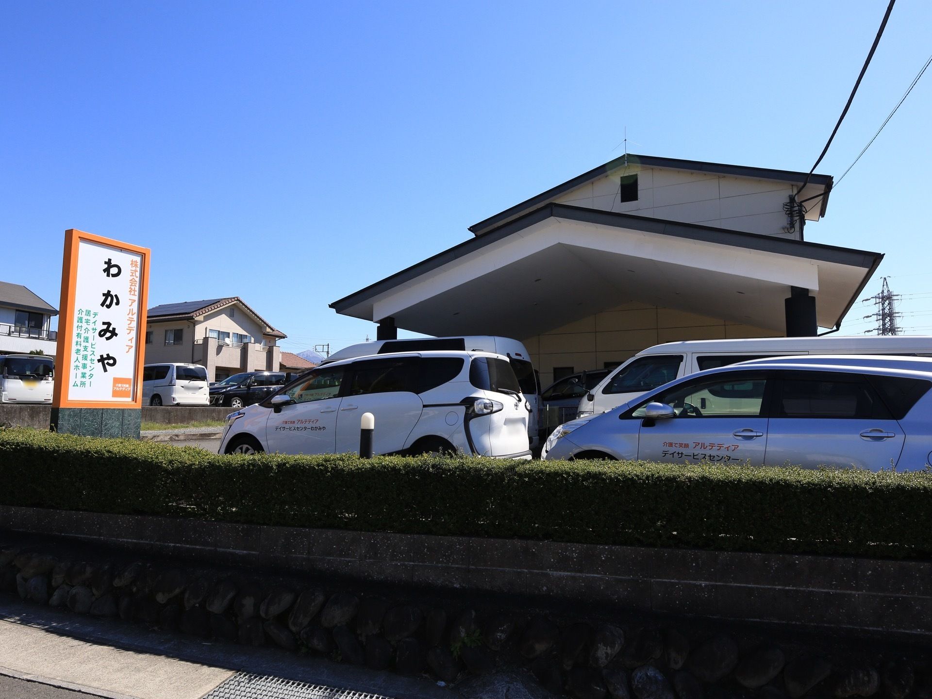 施設建物の前の植え込み沿いには、駐車場が完備され専用車が停車している。そばには外灯や施設の看板が立てられている。