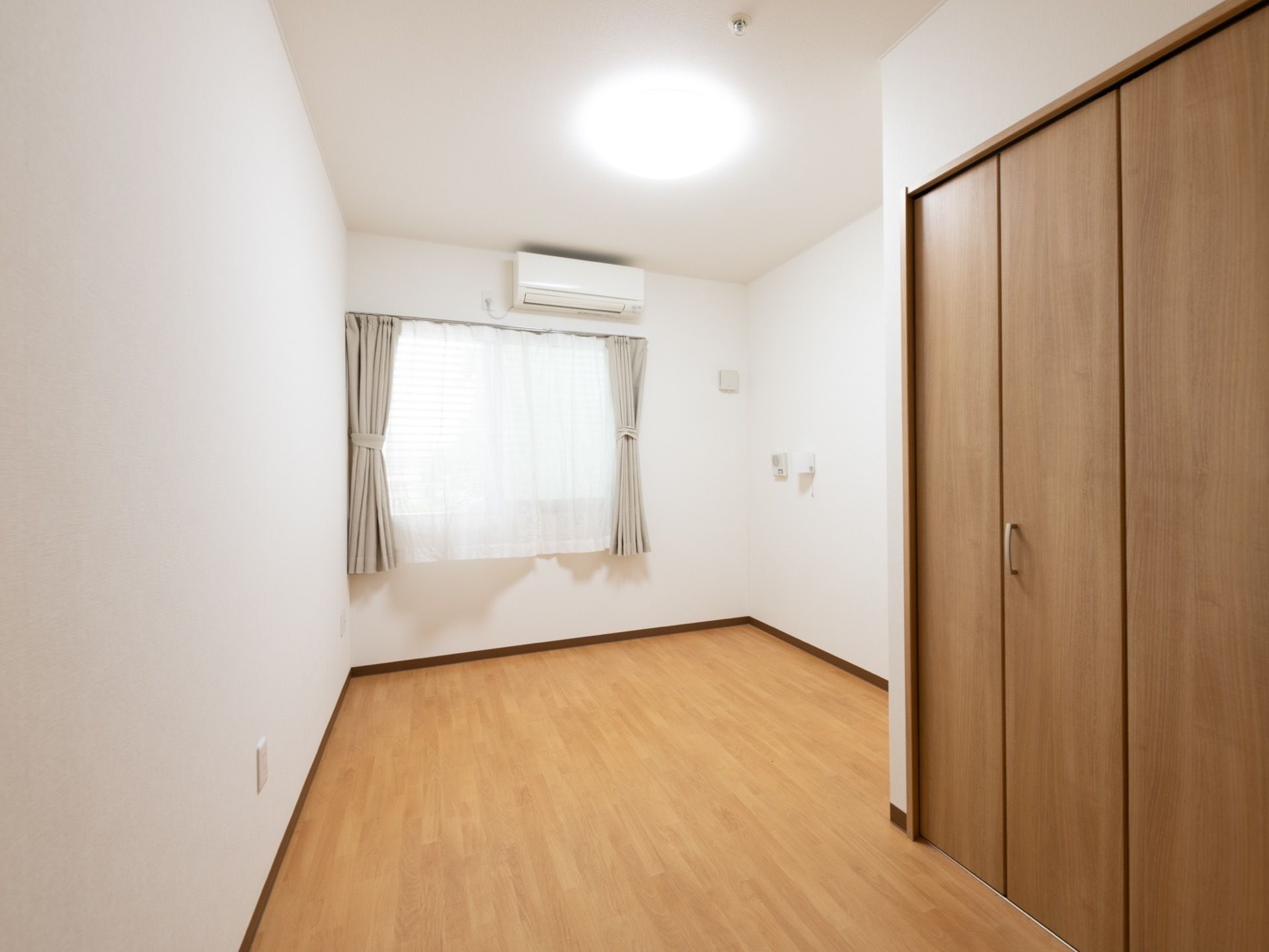 白い壁紙と木目調のフローリングのシンプルで明るい長方形の居室である。壁にはナースコールが設置されている。