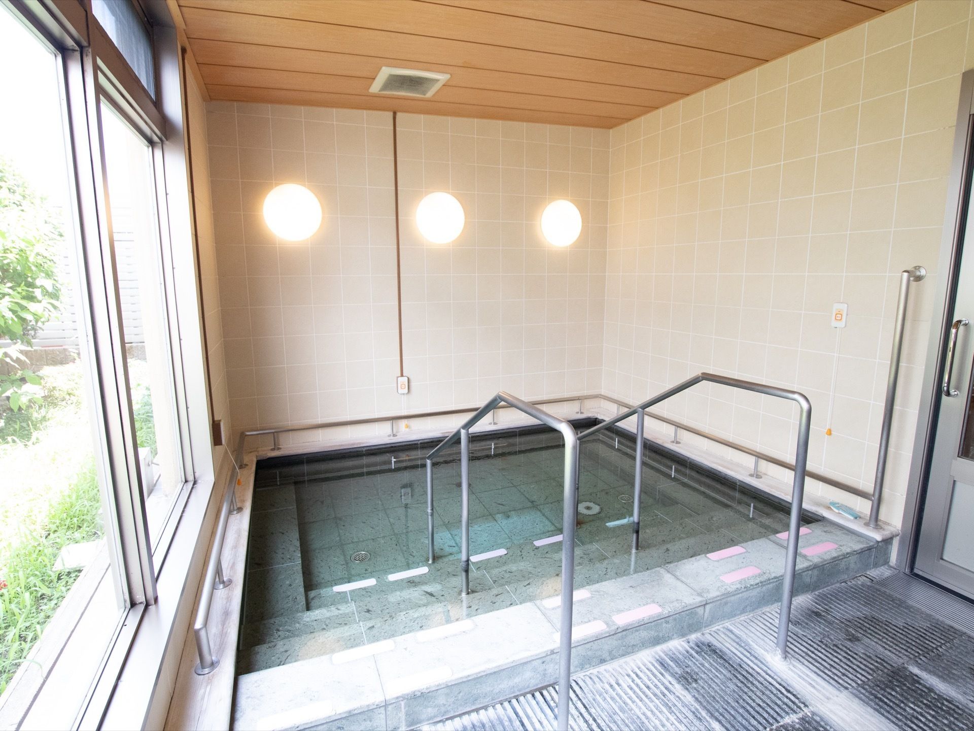 大きな窓のある大浴場である。壁に３つ並べて照明が取りつけられており、浴槽の周りや階段の両サイドには手すりが設置されている。
