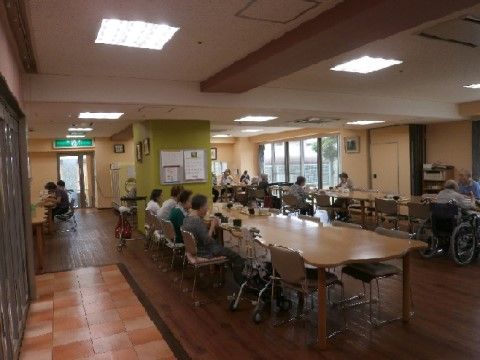 広くて明るい室内に、角が丸い長テーブルが３列並んでいる。テーブルには、車椅子で着席し、お食事をされる入居者様もおられる。