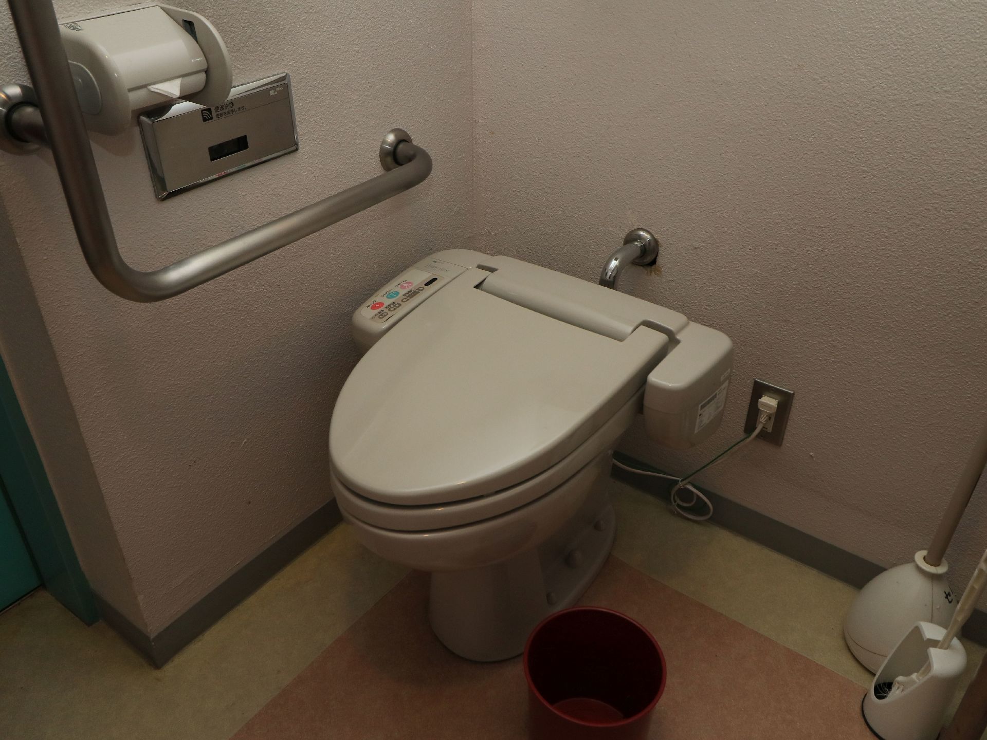 トイレには温水洗浄便座を設置している。壁には消音装置の他、固定式の手すりが取りつけられている。床に掃除用具が置かれている。