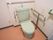 薄い緑色の洋式トイレがある。壁からステンレス製の手すりが出ている。手すりに緊急用ブザーがぶら下がっている。
