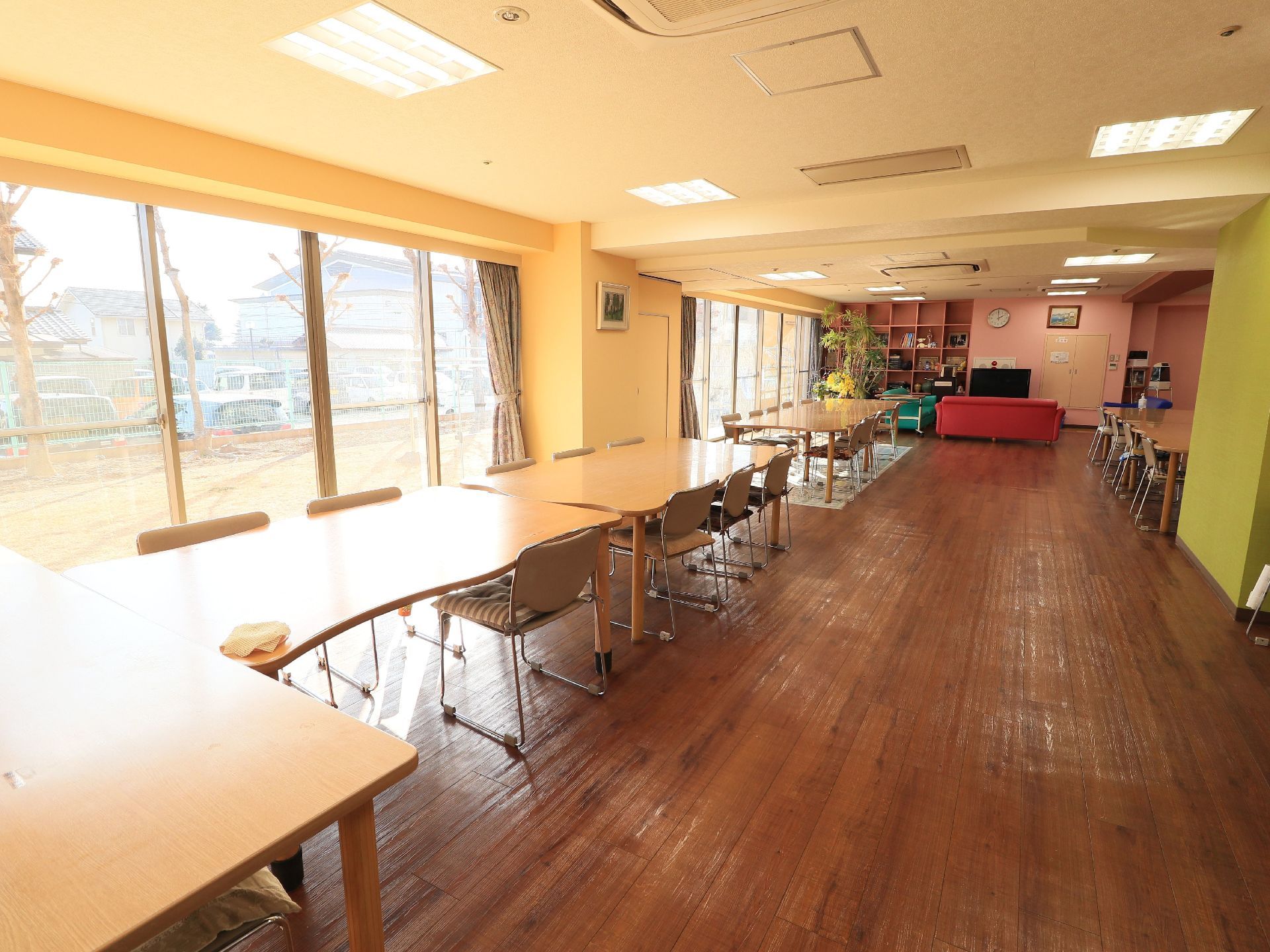 板張りで横に長い共有スペースで、窓が一面に開いている。カーテンが開けられ、外の景色が見えている。テーブルセットが長く置かれている。