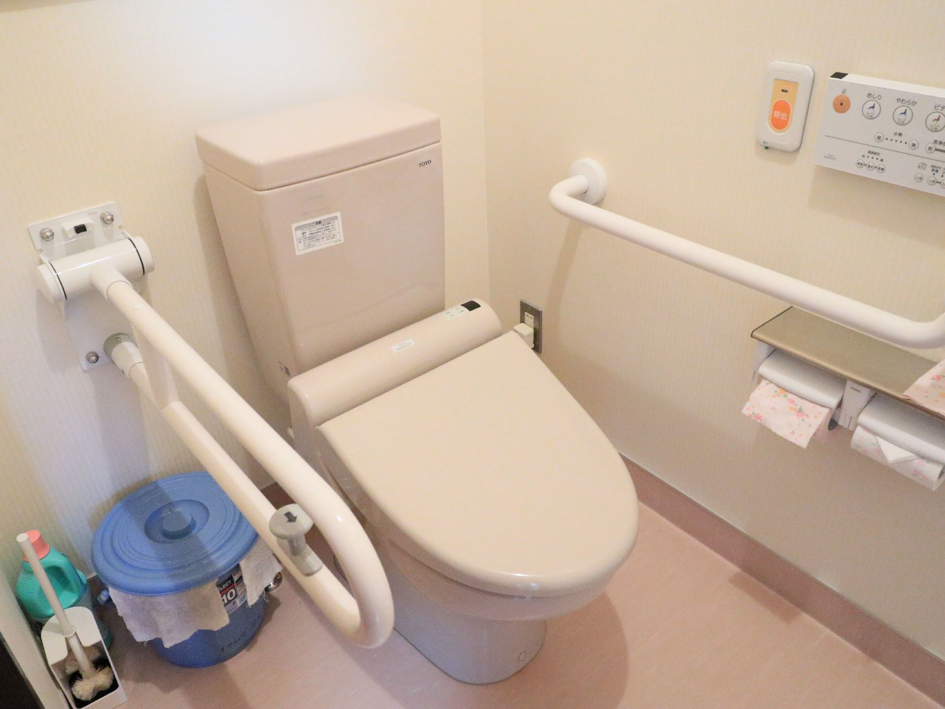 トイレはふたがついた便座になっている。便座に座って右側には、可動式のひじ掛けがある。右側の壁には横方向の手すりがついている。