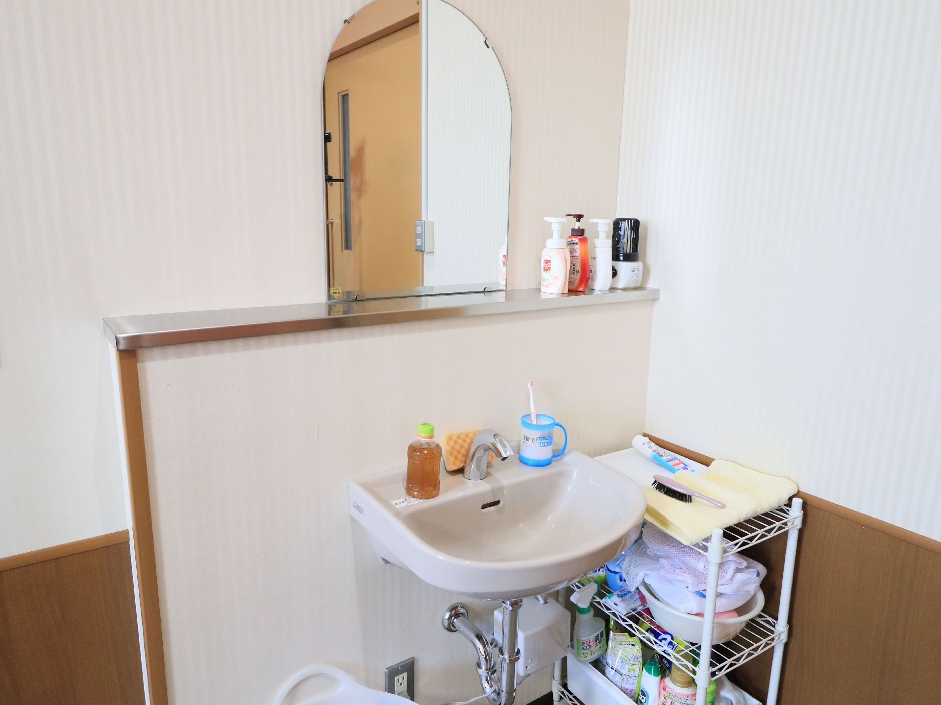 居室の入口付近に、洗面台が設置されている。小物を置く収納ラックが右側の壁際に置かれている。自動水栓になっている。