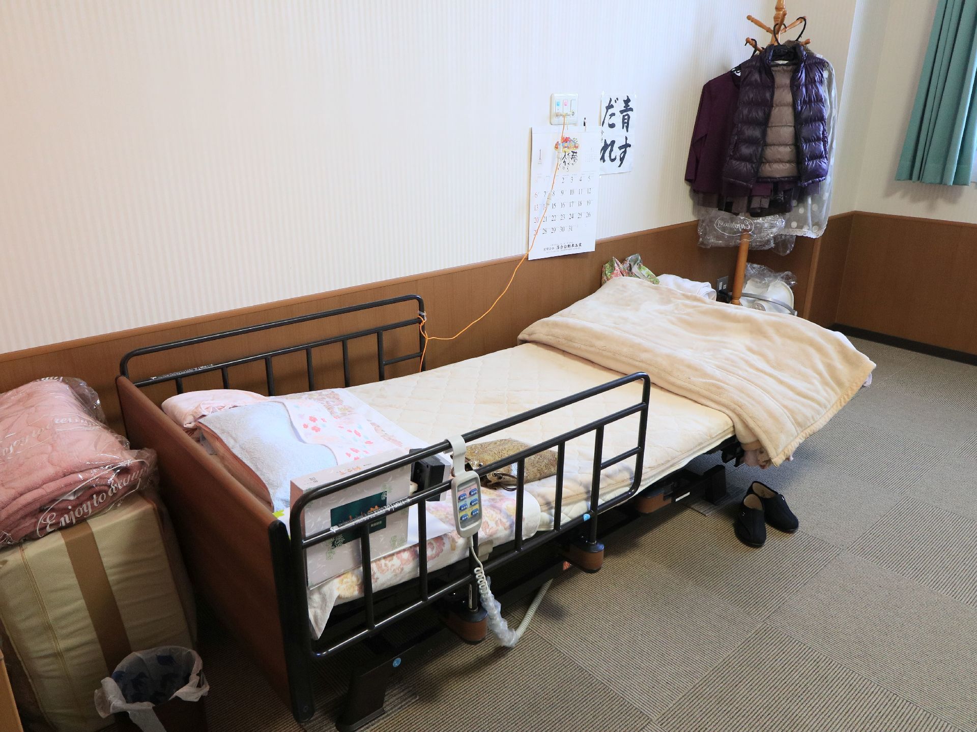 居室には、転落防止柵つきの介護ベッドが設置されている。ベッドの横の壁にはカレンダーや書道作品が貼られている。