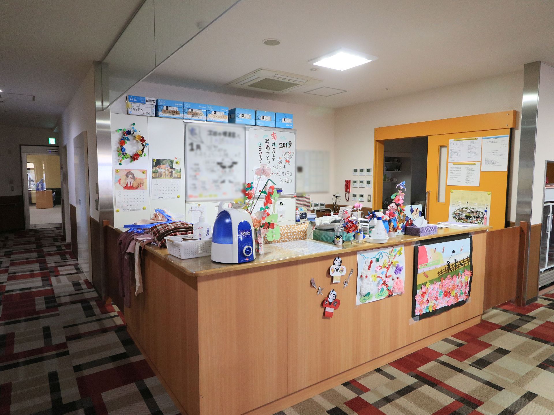 事務室前に受付がある。カウンターの上や前に、入居者様が作られたいろいろな作品が飾られている。時候の挨拶が壁に貼られている。
