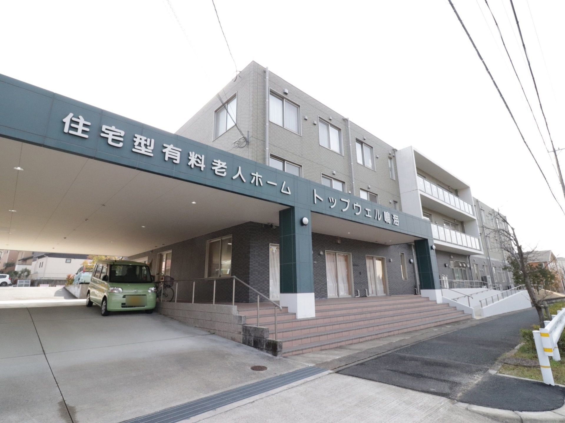 施設の外観の様子。3階建ての建物で、施設の入り口までは手すりの付いた階段が続いている。左はスロープで、駐車できる。