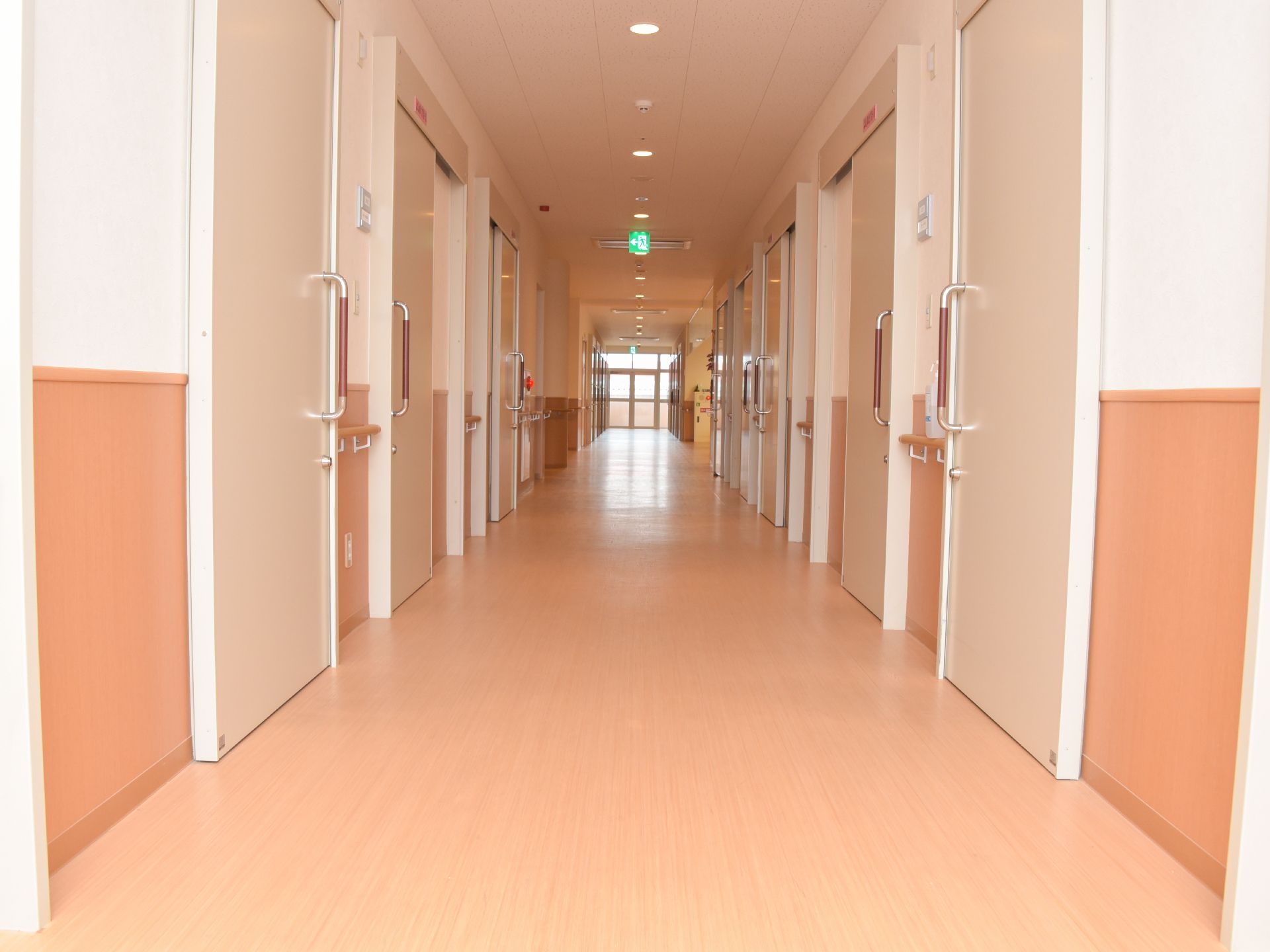 白色と木目仕様の廊下の内装である。両側には居室のベージュ色のドアが並んでいる。正面はガラス張りになっている。
