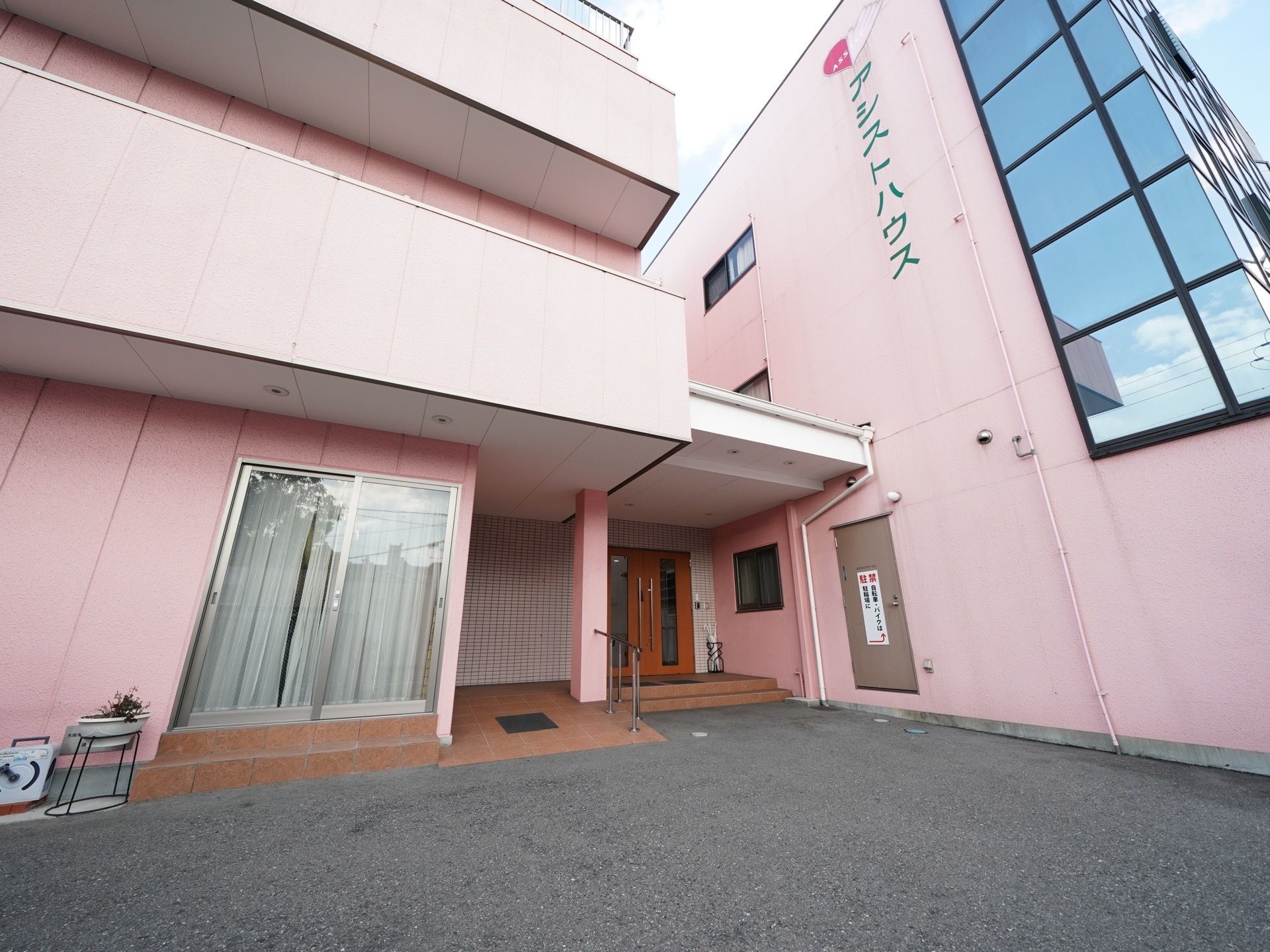 ピンク色の壁に大きく施設名が表示されている。建物は３階建てで入口前には階段とスロープが用意されている。
