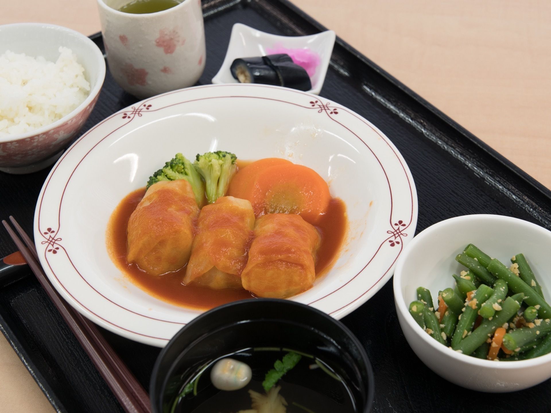 トマト系のソースがかかったロールキャベツと温野菜、インゲンの胡麻和えとご飯、すまし汁、香の物とお茶がお盆にセットされている。
