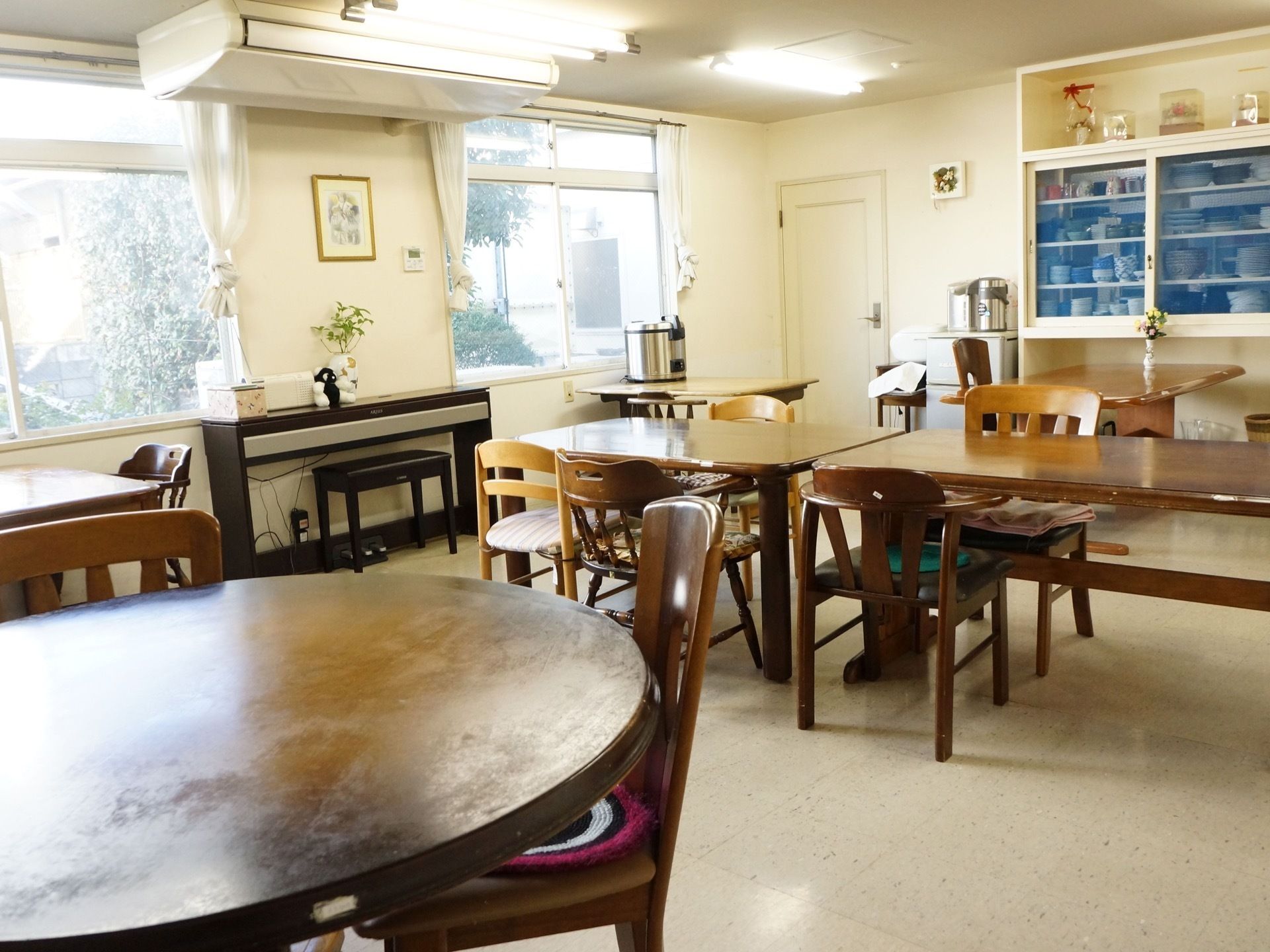 窓際のテーブルに炊飯器が置いてある。部屋の奥に食器棚があり、その横に冷蔵庫が置いてある。そのそばに白い扉がある。