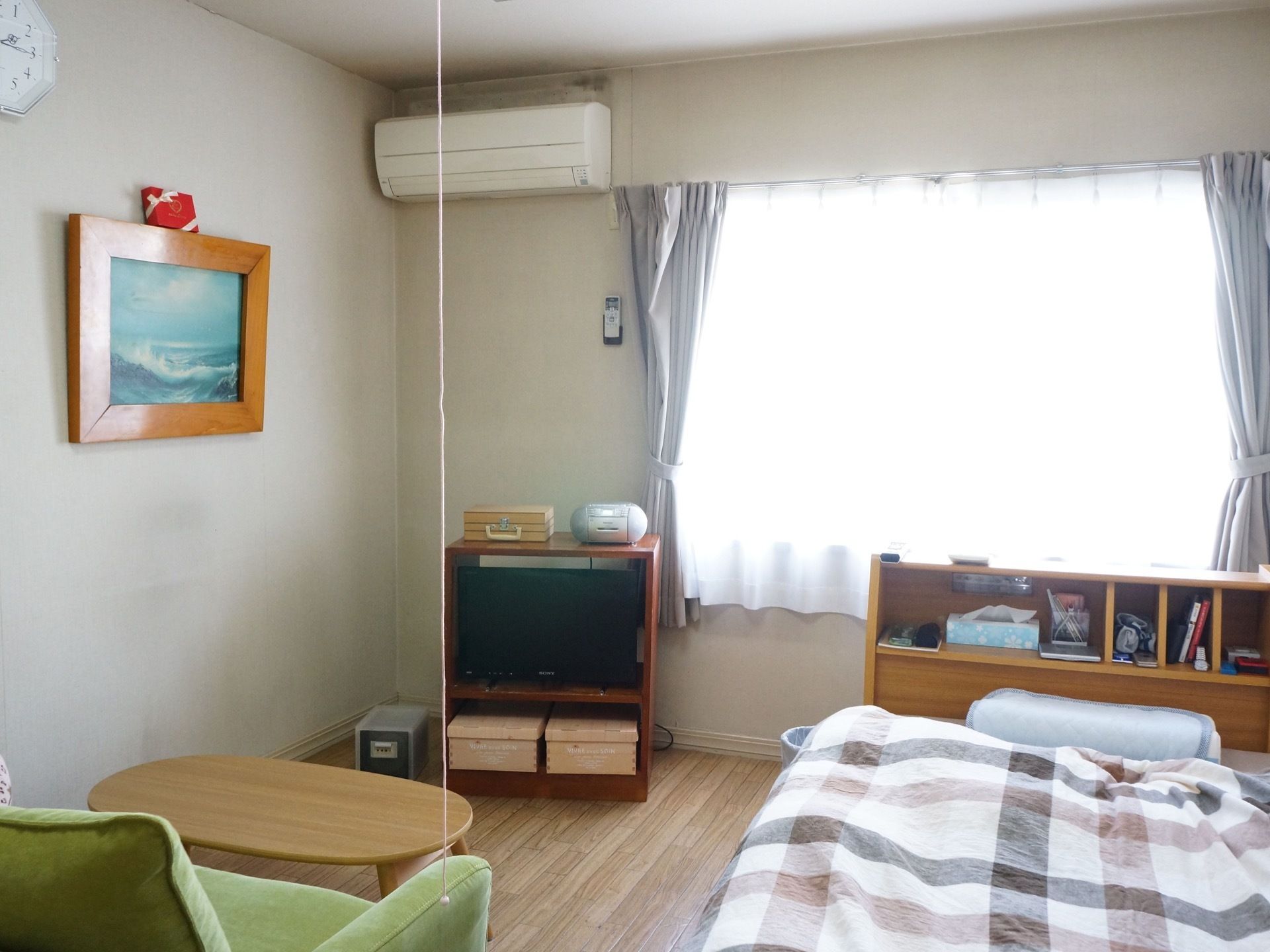 ベッド、テレビ、テーブル、椅子が置いてある。窓があり、カーテンが二重につけられている。壁に絵が飾られている。