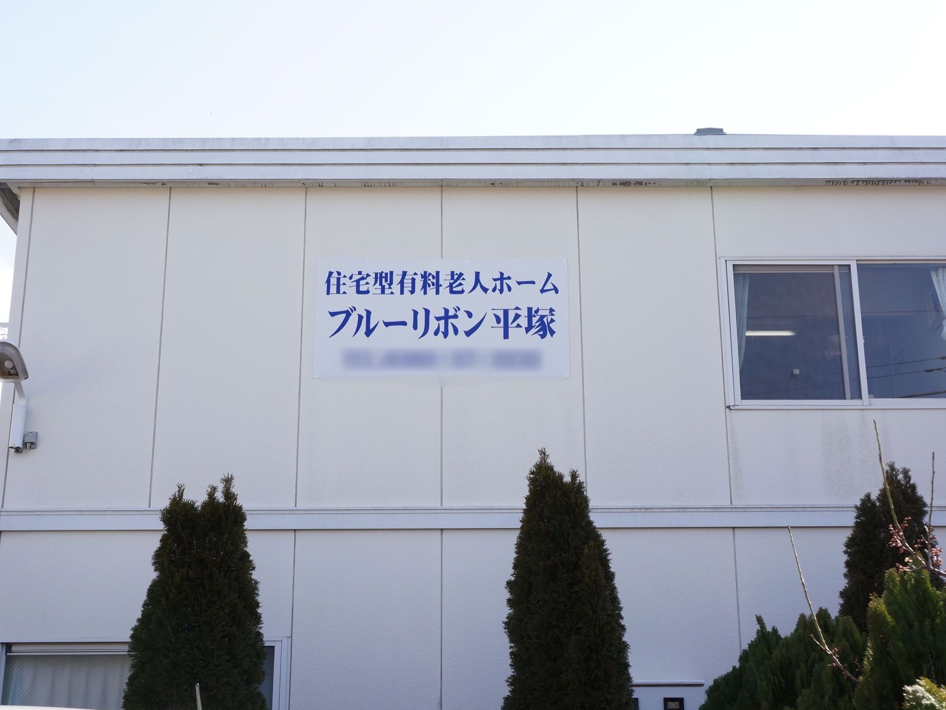 白い壁の２階建ての建物である。壁に施設名が掲げられている。その横に窓がある。建物のそばに木々が何本も植えられている。