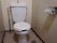 白い便座とタンクが設置されている。壁にトイレットペーパーが設置されている。床と両側の壁はベージュ色である。