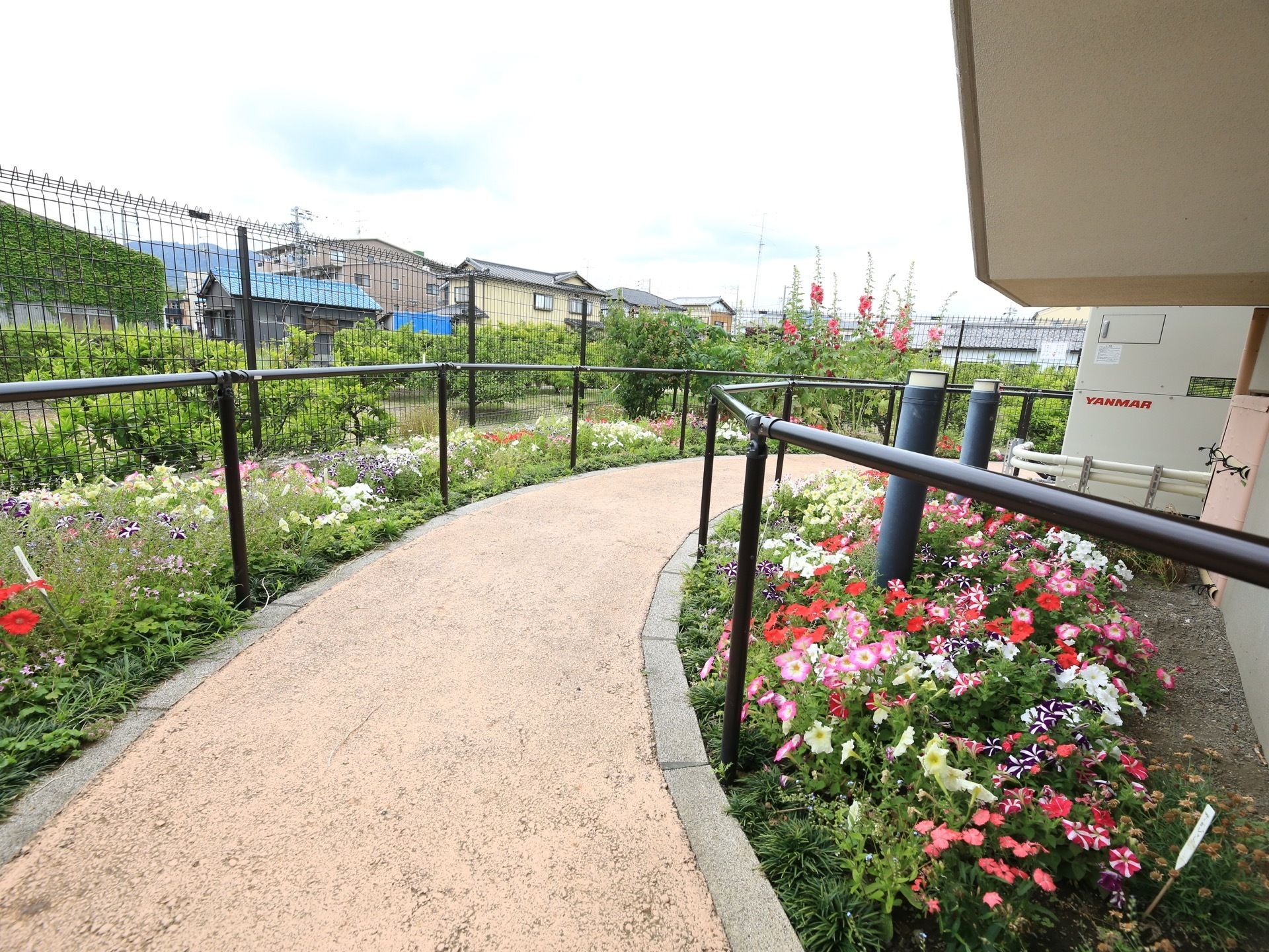 敷地内に、自立式の手すりを備えた遊歩道が設置されている。両側に花壇があり、色とりどりの花が咲き誇っている。