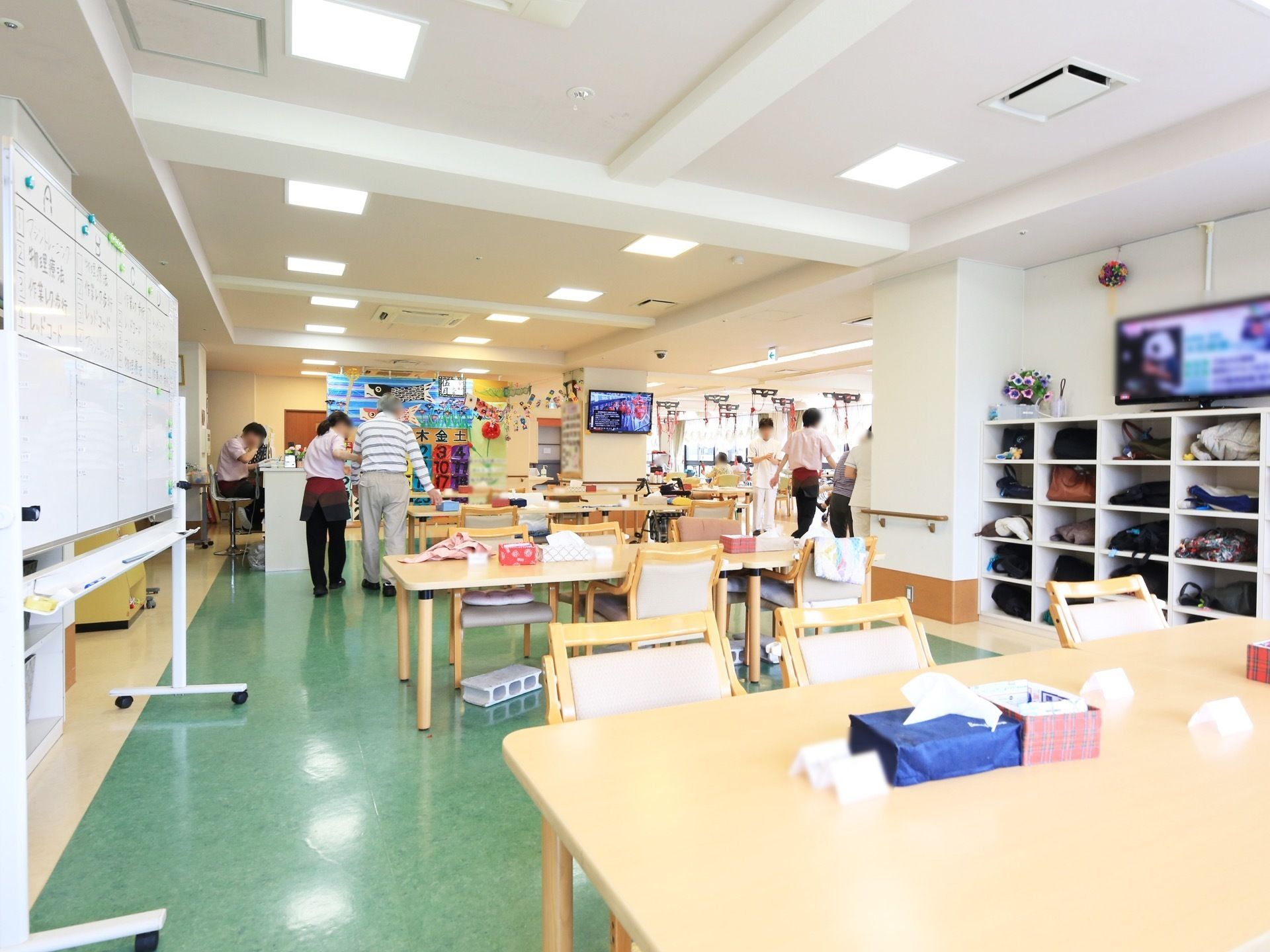 多くのテーブルや椅子が置かれた共有スペースがある。男性の入居者様が、スタッフの手を借りながら歩いている。