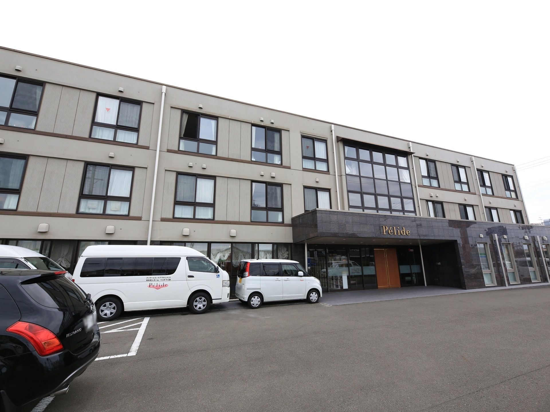 薄い灰色を基調とした外壁の３階建ての施設である。玄関前には広いスペースがあり、車が何台も停まっている。