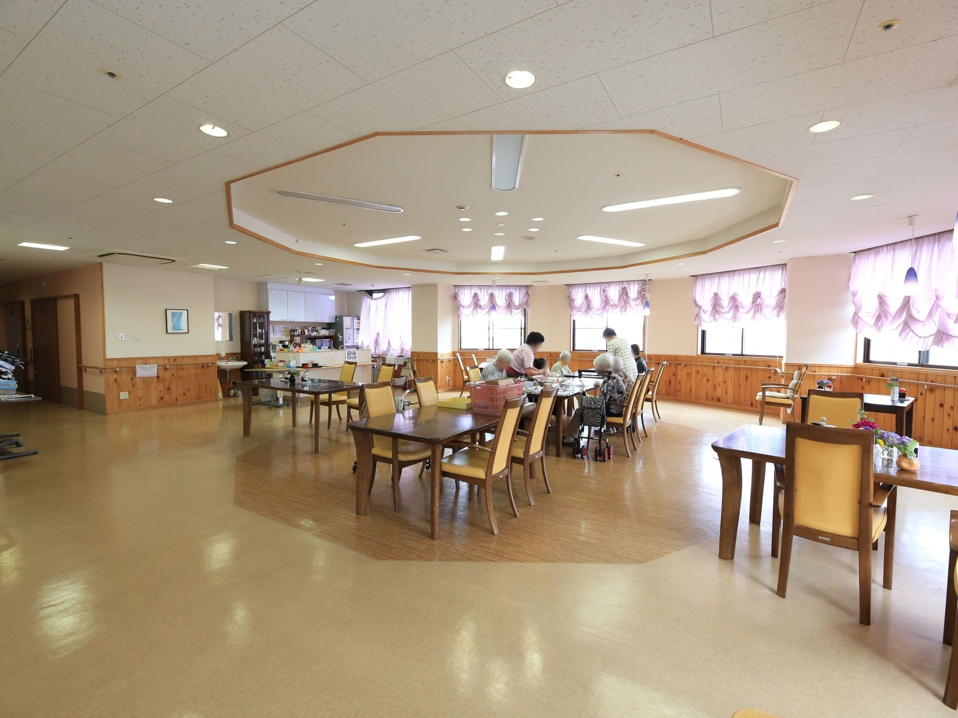 半円形状に張り出した空間が食堂になっている。複数の入居者様がテーブルについていて、スタッフが２名作業をしている。