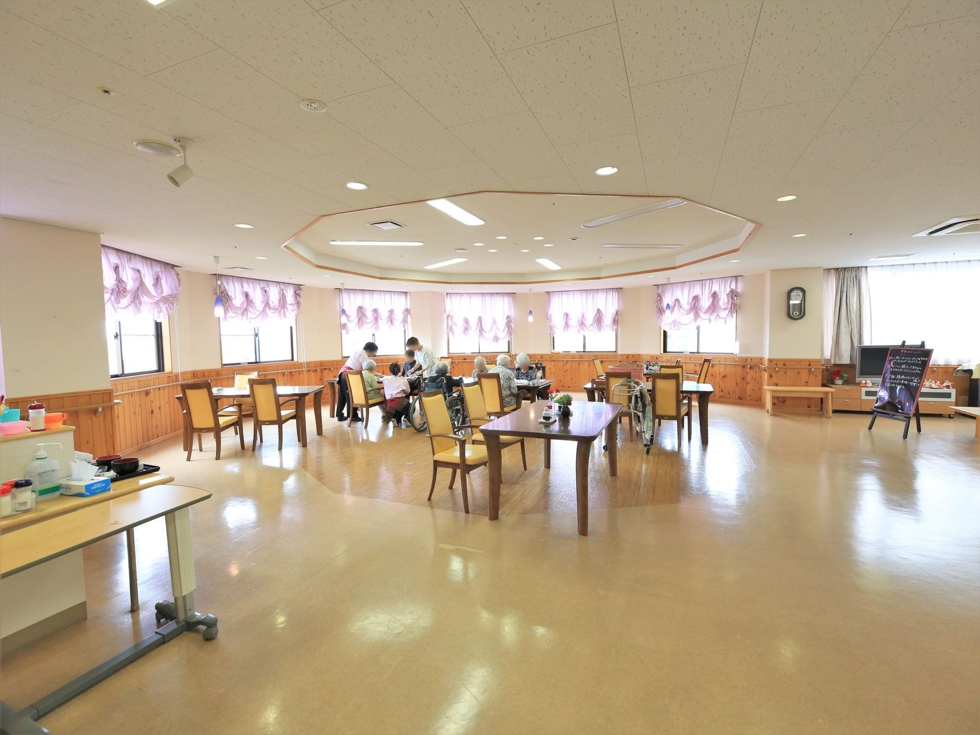 食堂にある２つのテーブルに入居者様が何名か集まっている。その近くで、女性スタッフ２名が作業をしている。