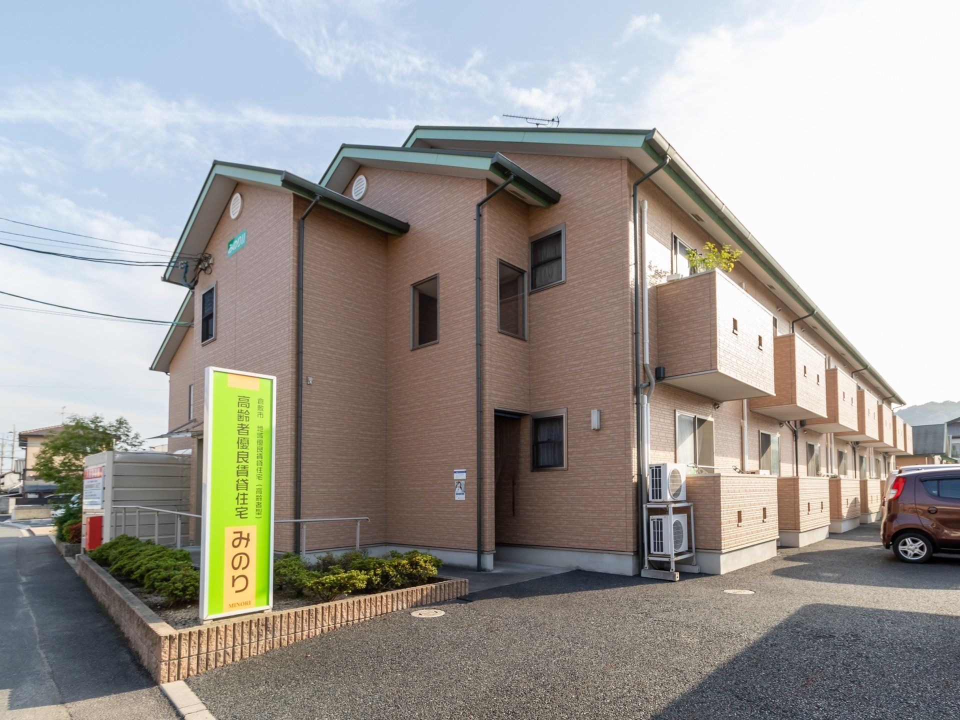 ブラウンを基調とした２階建ての建物で、各部屋にはベランダが設けられている。施設入口には、看板が設置されている。