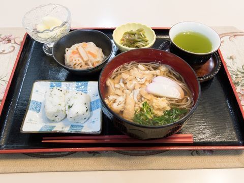 ランチョンマットの上に、お盆、その上に食事が載っている。角皿におにぎり、そばのほか、丸型の小鉢が2点ある。フルーツとお茶もついている。