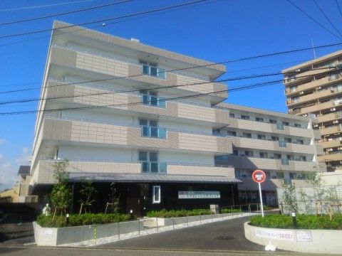 ベージュの５階建て新築の建物は、２階から上の階にバルコニーがある。敷地内には手入れが行き届いた植栽がある。