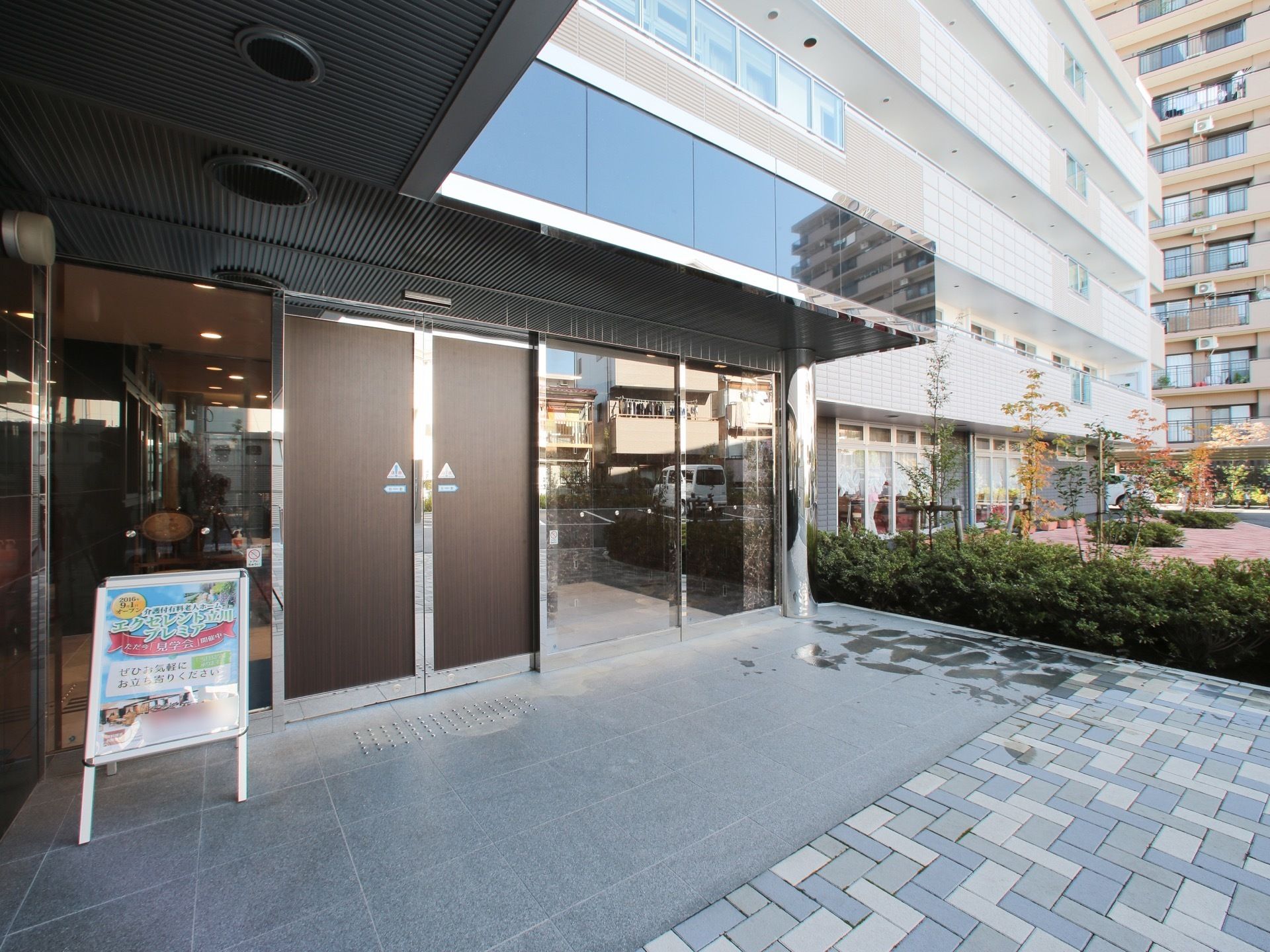 施設の入口部分は広いスペースがあり、雨除けの屋根もある。右手には植物がたくさん植えられていて、自動ドア付近には看板もある。