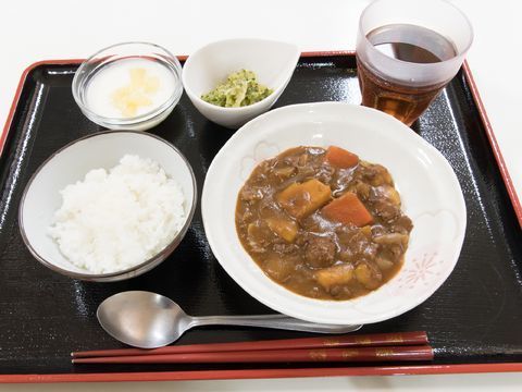 黒いお盆の上に食事が乗っています。カレーとごはんが別皿に盛り付けてある。スプーンがある。ガラスの器と小鉢がついている。