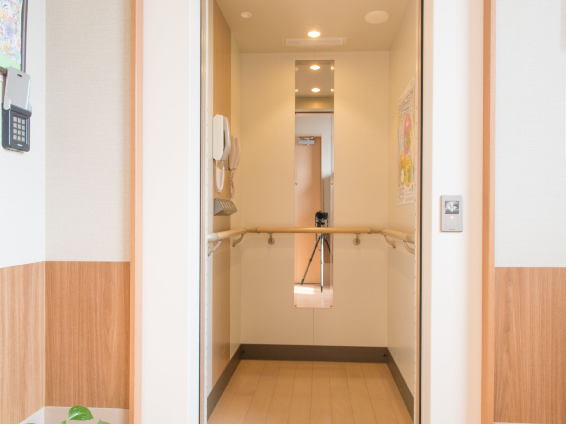 エレベーター内部には、電話などが壁に取り付けてある。ダウンライトが天井にある。手すりがついている。フローリングになっている。