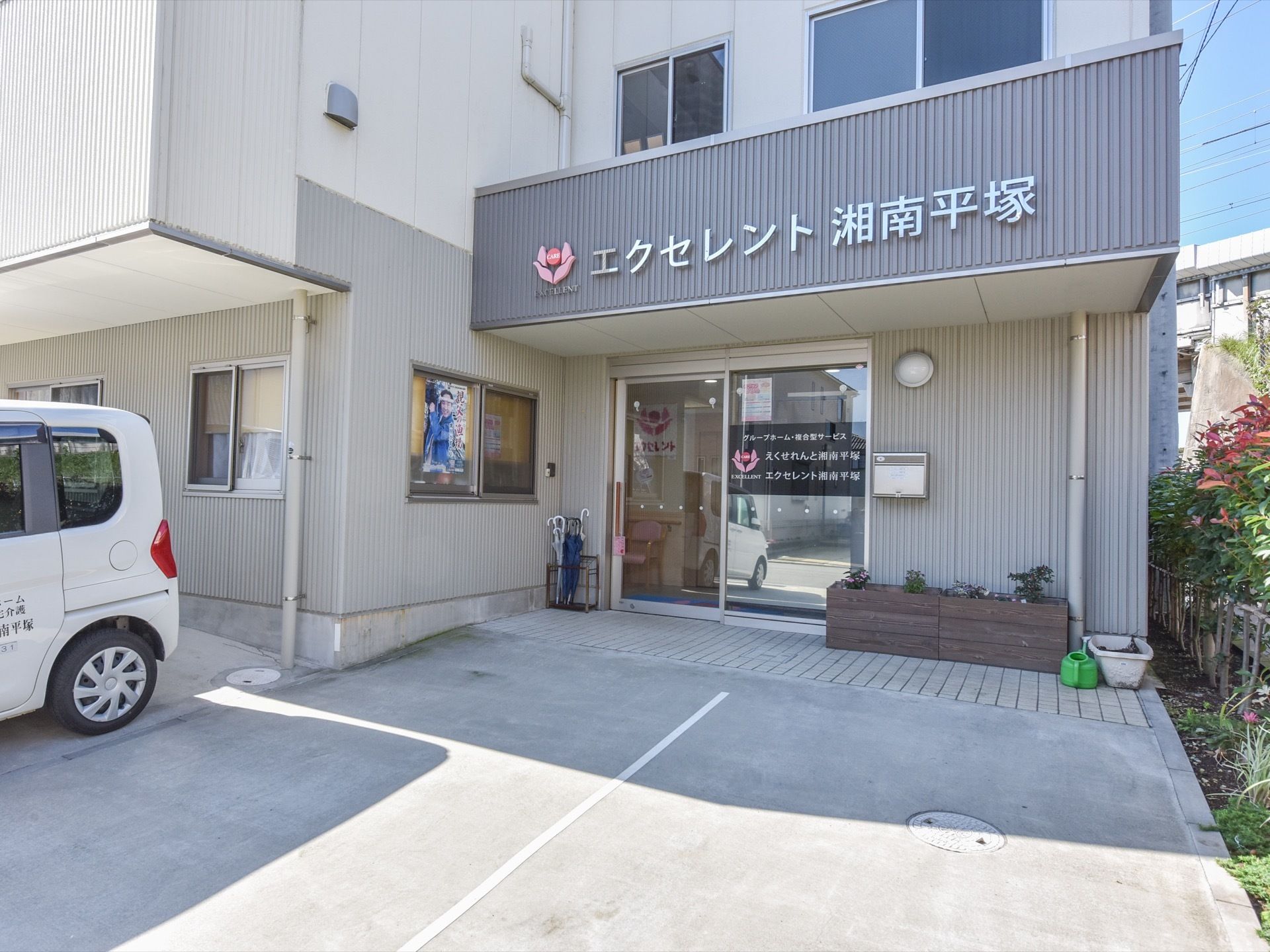 玄関には、大きくて透明な扉がある。施設の名前がとても大きく表示されている。外には傘立てが置かれている。